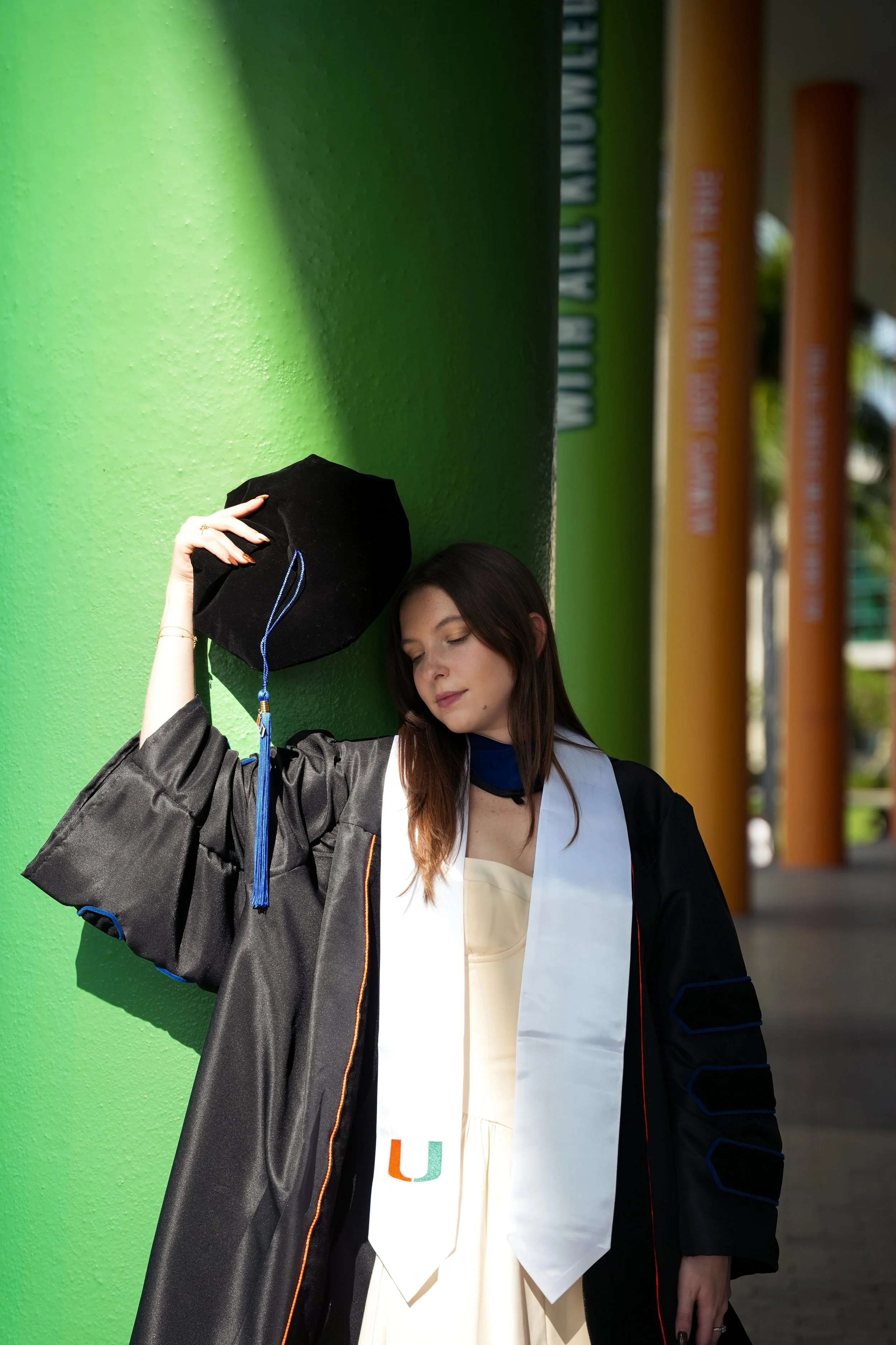 A young woman in a graduation gown and cap holding her cap in her hand, leaning against a bright green wall with her eyes closed.