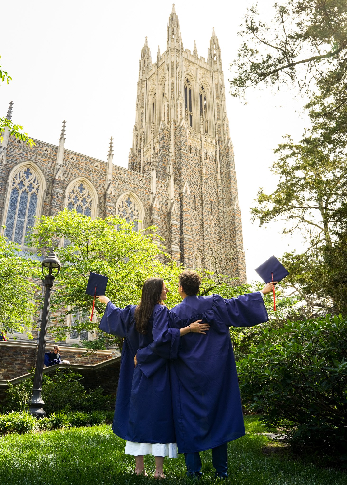 Two graduates in blue gowns celebrating outdoors on graduation day in front of a tall, ornate church with gothic architecture and large stained glass windows. The woman is hugging the man, who has his arm around her waist, and they're holding graduat