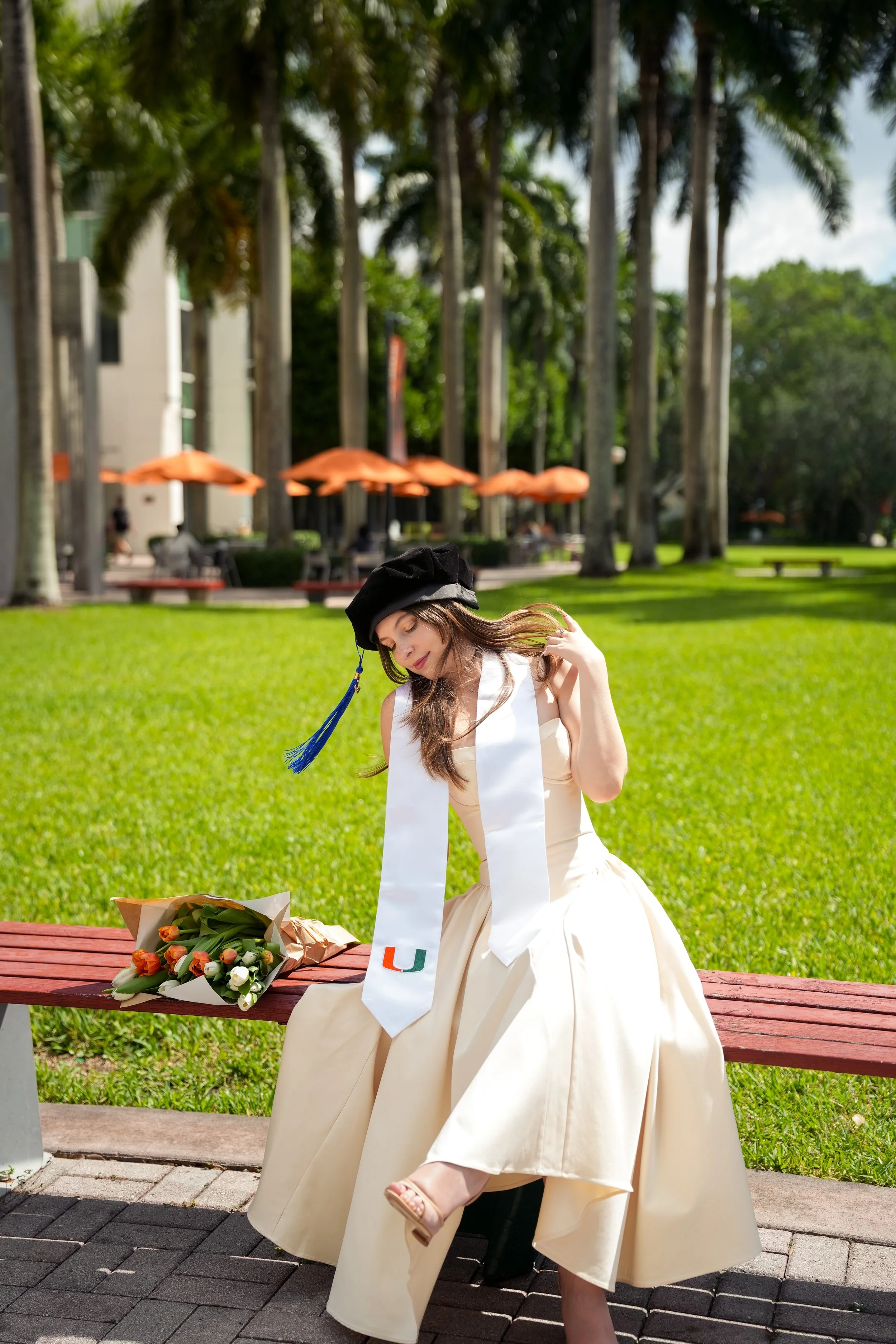 A young woman in a white graduation gown and black cap with a blue tassel sits on a wooden bench outdoors on a sunny day. She is holding some of her long hair with her right hand. In front of her on the bench is a bouquet of flowers, and around her n