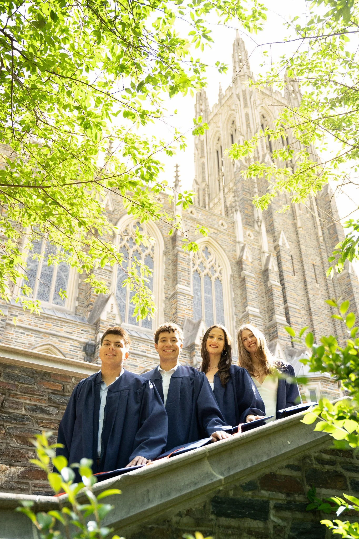 Four young graduates in caps and gowns standing on a stone staircase outside a historic stone church or cathedral with tall windows and gothic architecture, framed by leafy green trees.