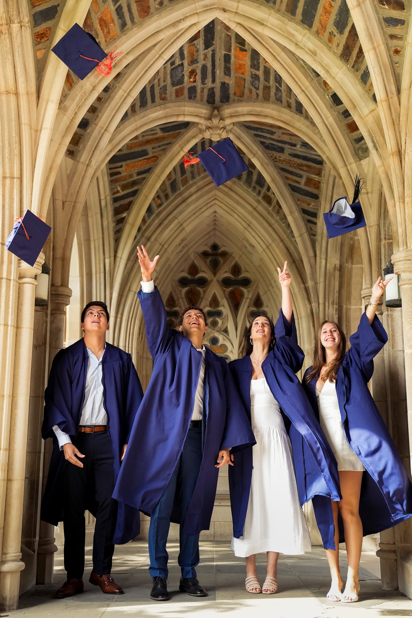 Five graduates in blue gowns and caps celebrating in a historic stone hall, tossing their caps in the air and smiling, with an arched ceiling and decorative architectural details in the background.