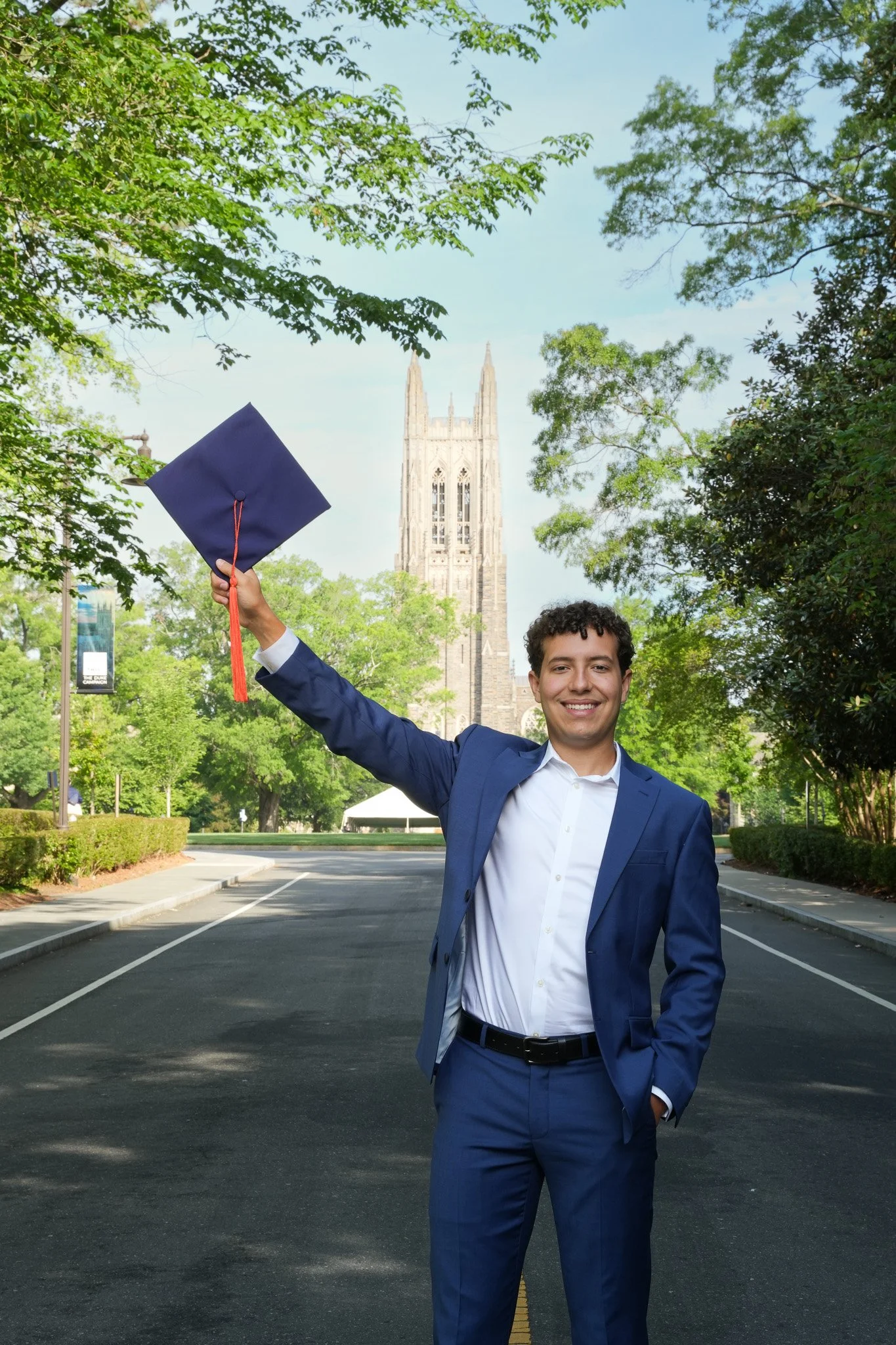 Young man in a blue suit holding a graduation cap in front of a church in a park with green trees.
