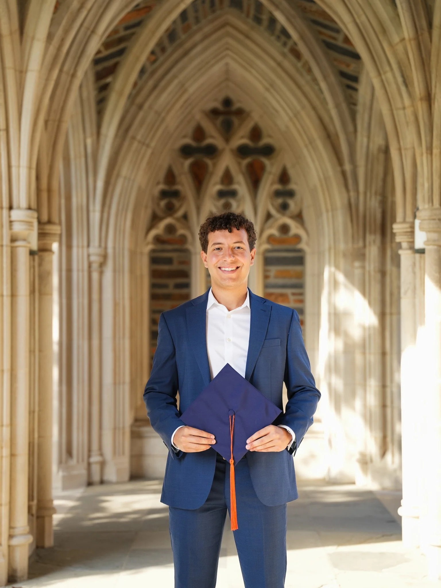 A young man in a blue suit and white shirt smiling while holding a graduation cap, standing in a cathedral-like hallway with arched ceilings and stained glass windows.