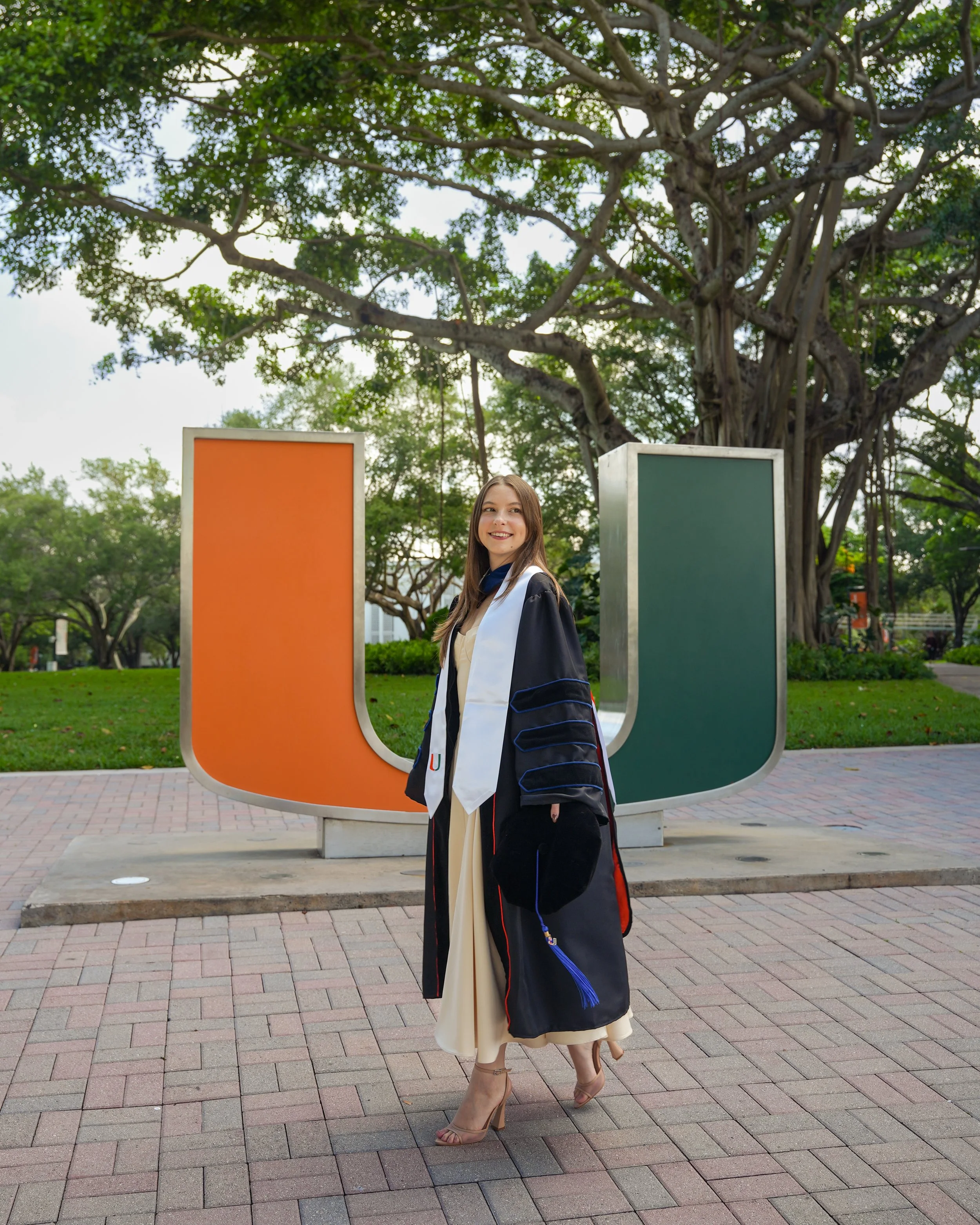 Young woman in graduation gown and cap standing outdoors in front of a large U-shaped sign, with a big tree and green foliage in the background.