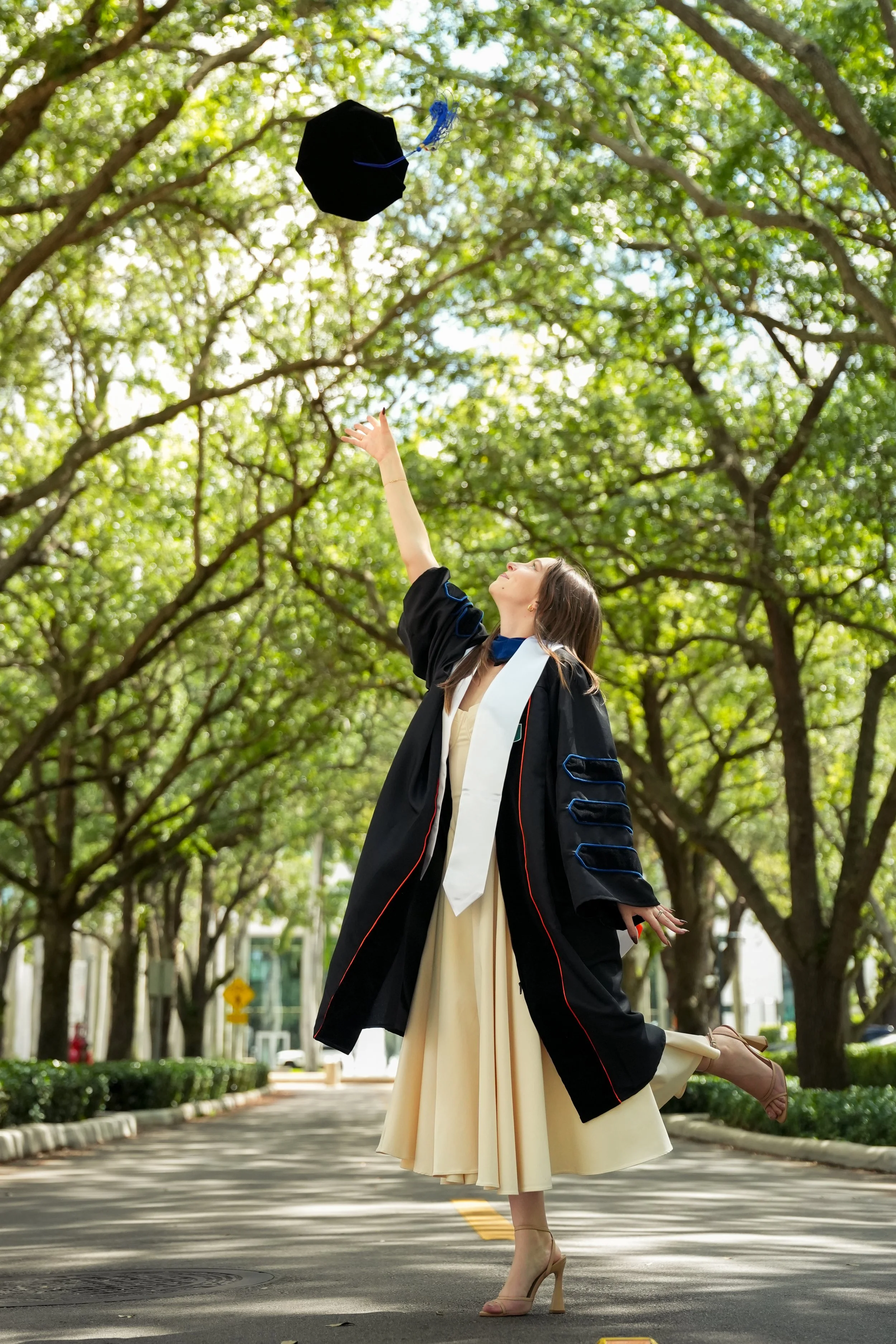 A woman in a graduation gown and cap stretching her leg and reaching up towards her cap as it is flying away in the air, standing on a street lined with trees.