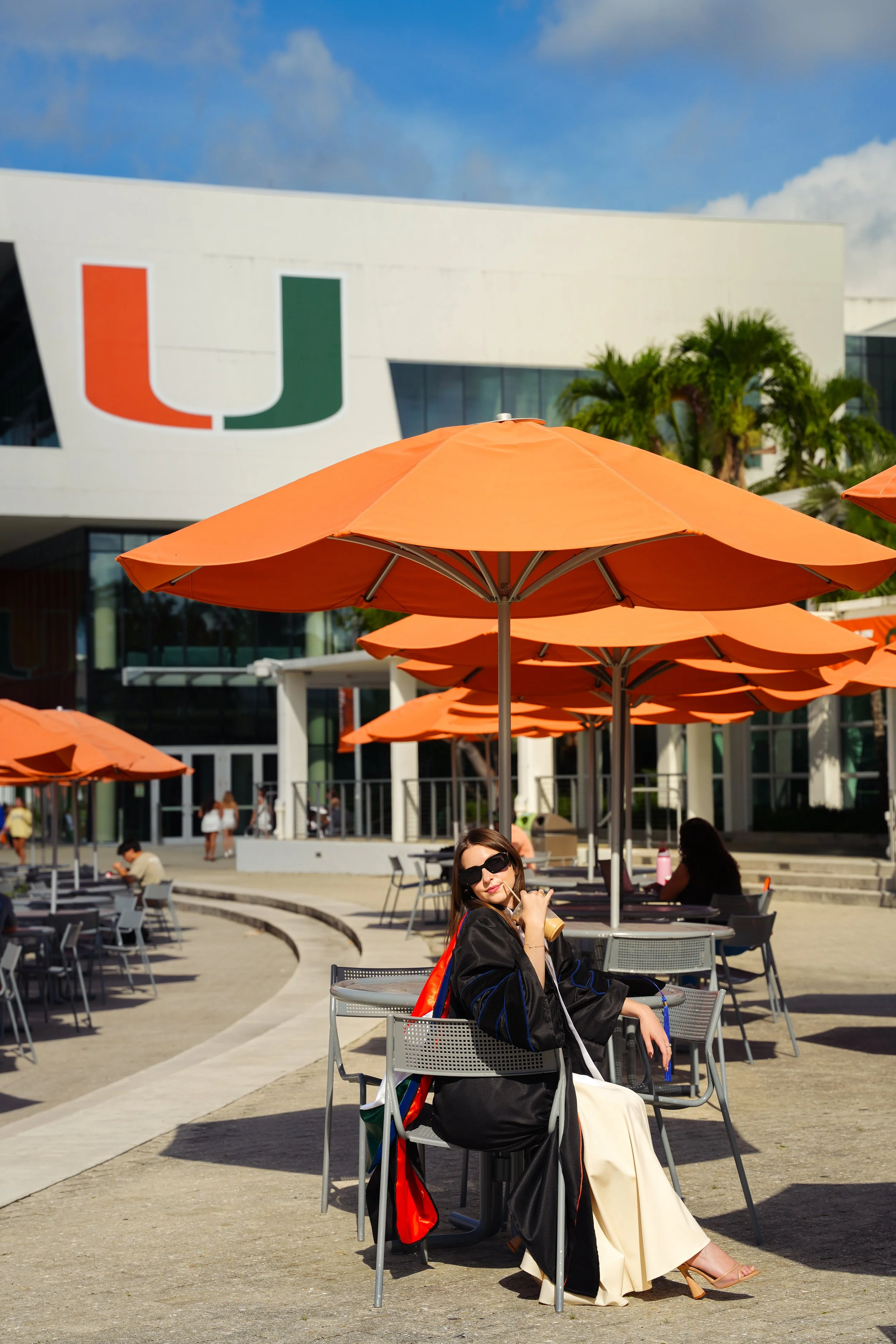 A woman sitting at an outdoor table with orange umbrellas, wearing sunglasses and fashionable clothing, at a university campus with the University of Miami logo on a modern building in the background.