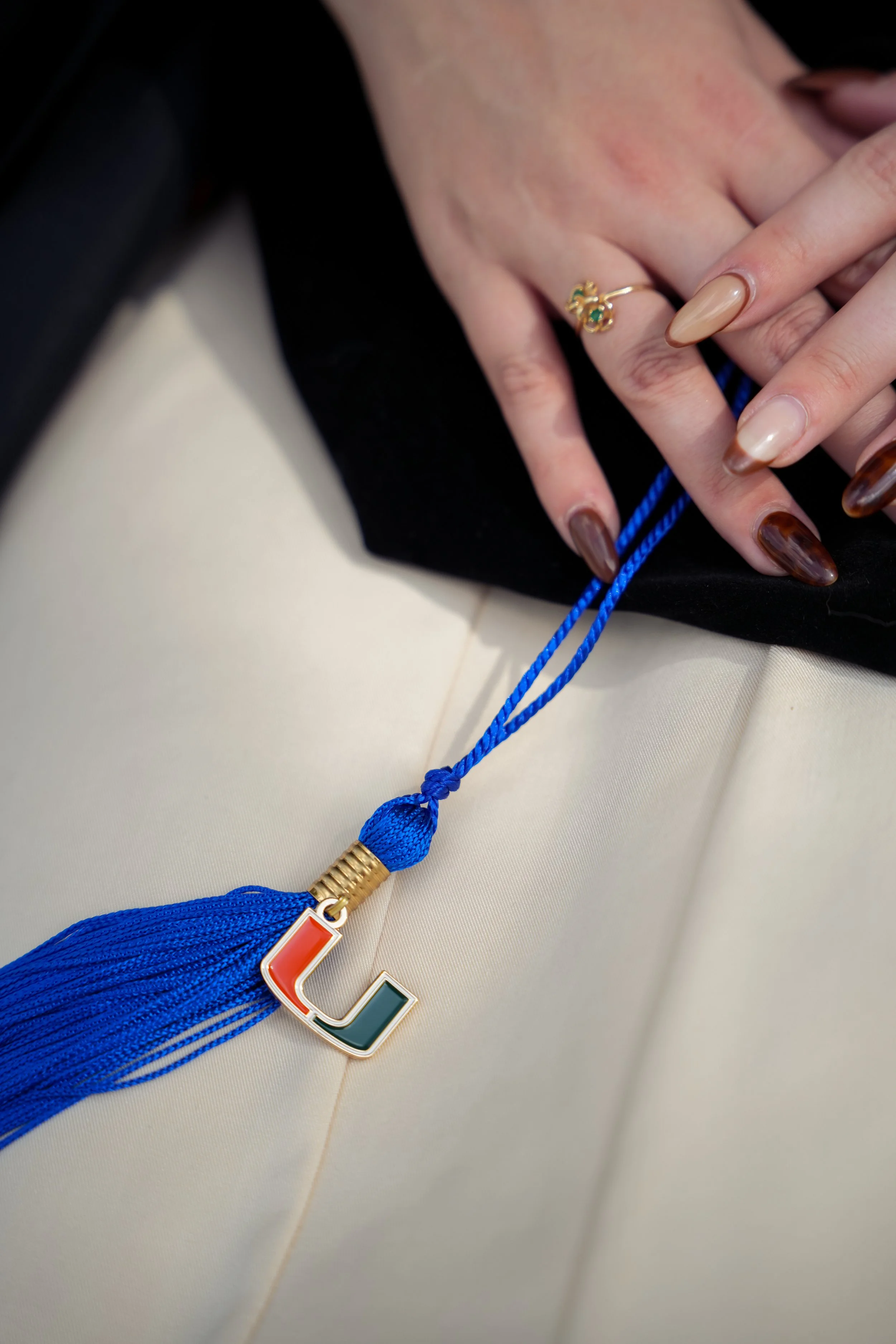 Close-up of a person's hand with manicured nails holding a blue tassel with a logo charm. The person is wearing rings, including a gold ring with green stones, and is dressed in a black and beige outfit.