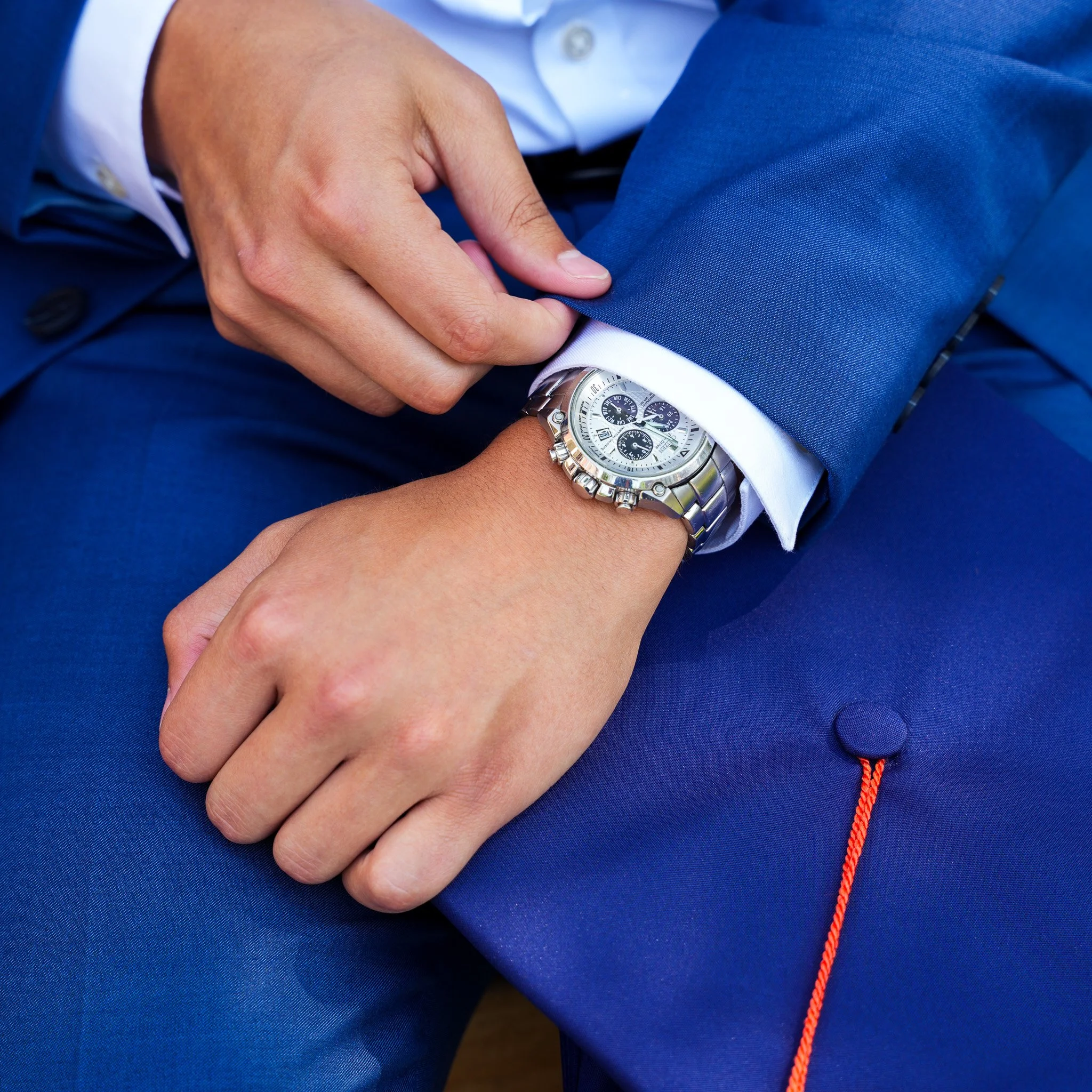 A man in a blue suit and white shirt is sitting with his left hand resting on his lap. He is wearing a silver wristwatch with a white dial and multiple subdials. The man is also wearing a black belt with a silver buckle and has a red string attached 
