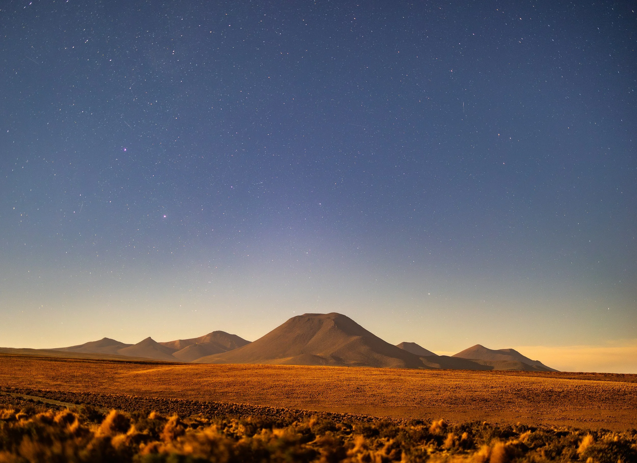 Nighttime landscape of a mountain range with a starry sky, some shooting stars, and a small crescent moon on the horizon.