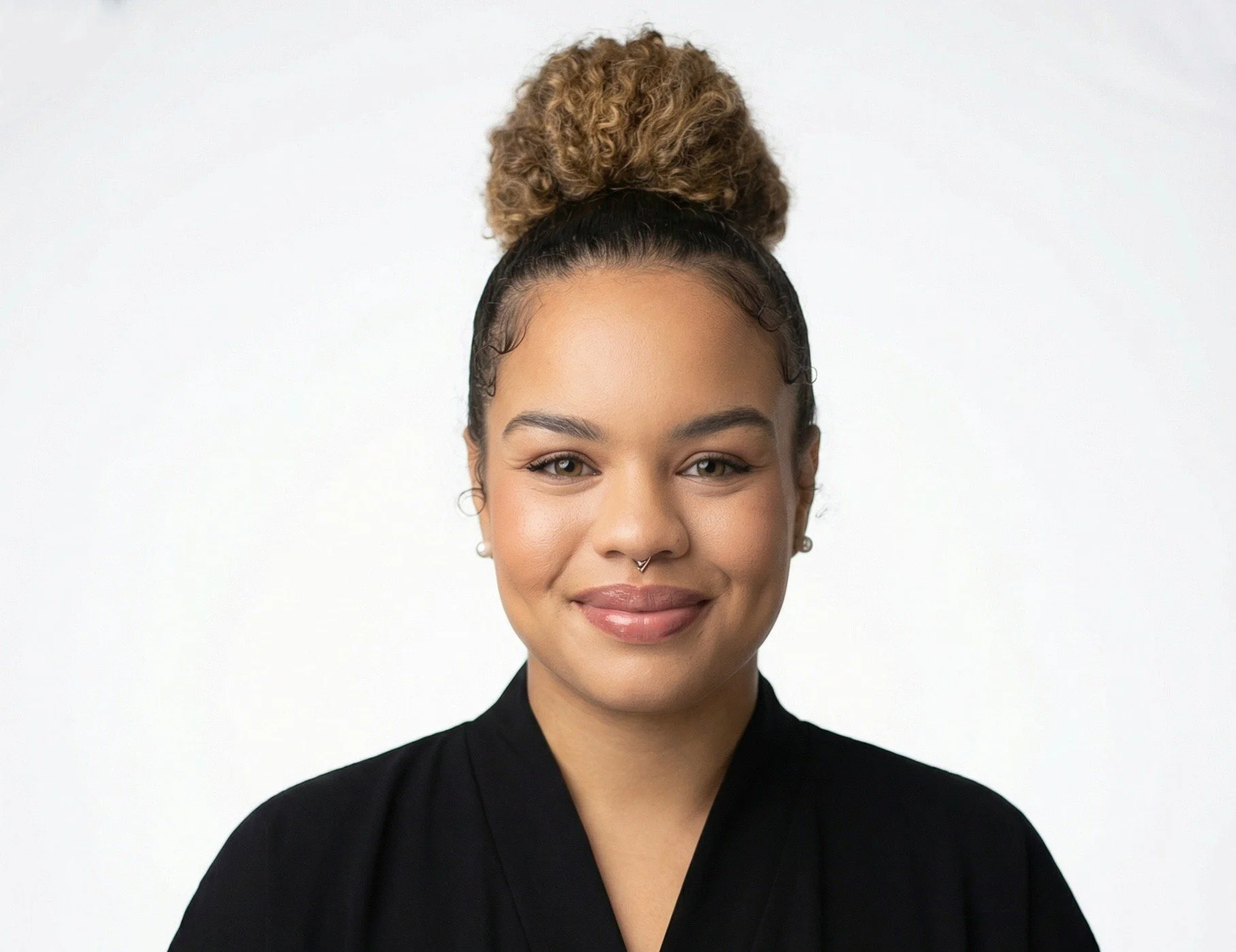 Headshot of a woman with light brown skin, curly blonde hair in a bun, wearing a black top, smiling, against a plain white background.