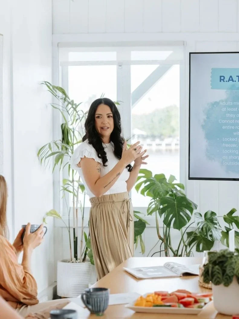 Candice Heffernan giving a presentation for an Amity Behaviour client in a bright room with large windows, plants, and a screen displaying information.