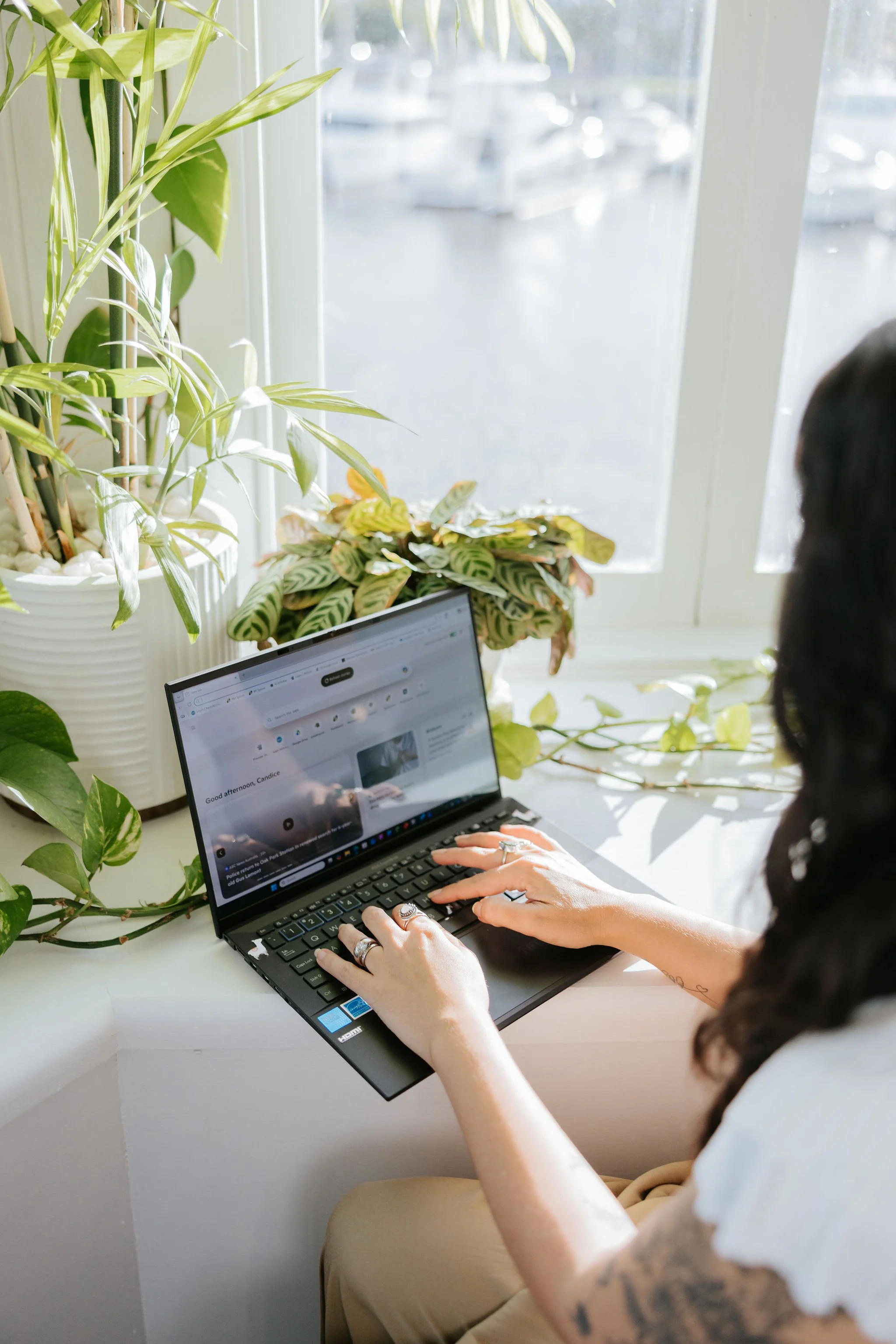 Candice Heffernan working on her laptop. She is sitting at a white table near large window with plants on the windowsill. Sunlight is streaming in, illuminating the scene.