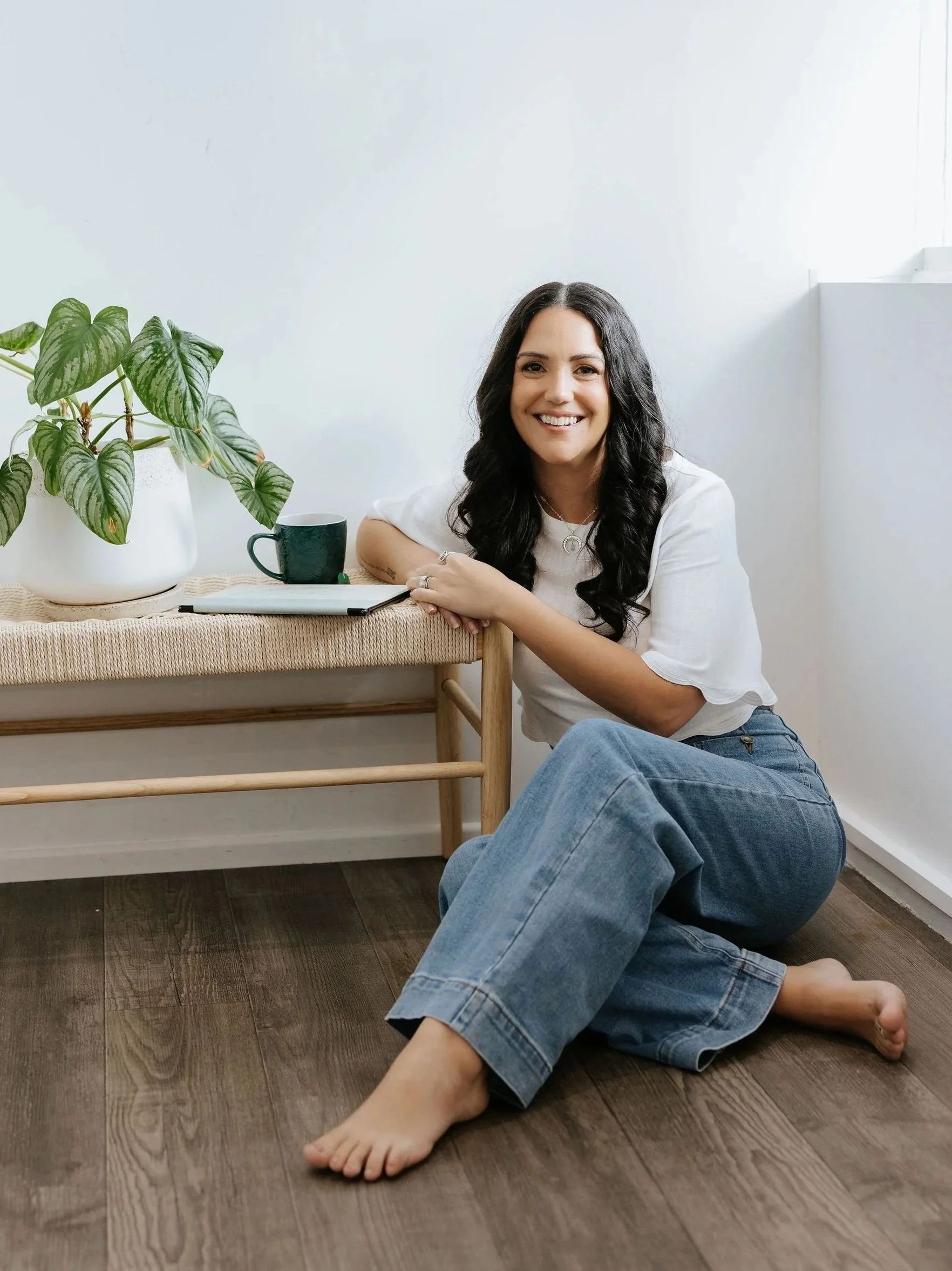 Candice Heffernan smiling while sitting on a dark hardwood floor next to a wooden bench, on which there is a white pot with green leafy plant, a black mug, and a closed notebook.