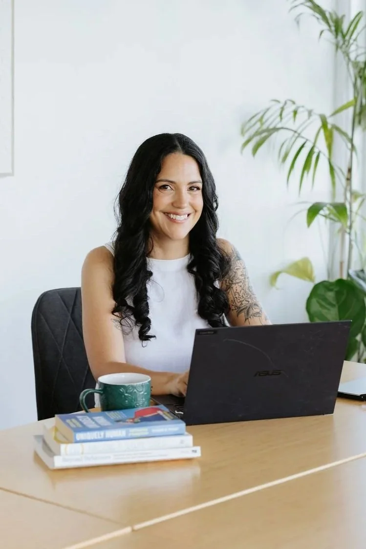 Candice Heffernan sitting at a desk with a laptop, books, a coffee mug, and greenery in the background, smiling at the camera.