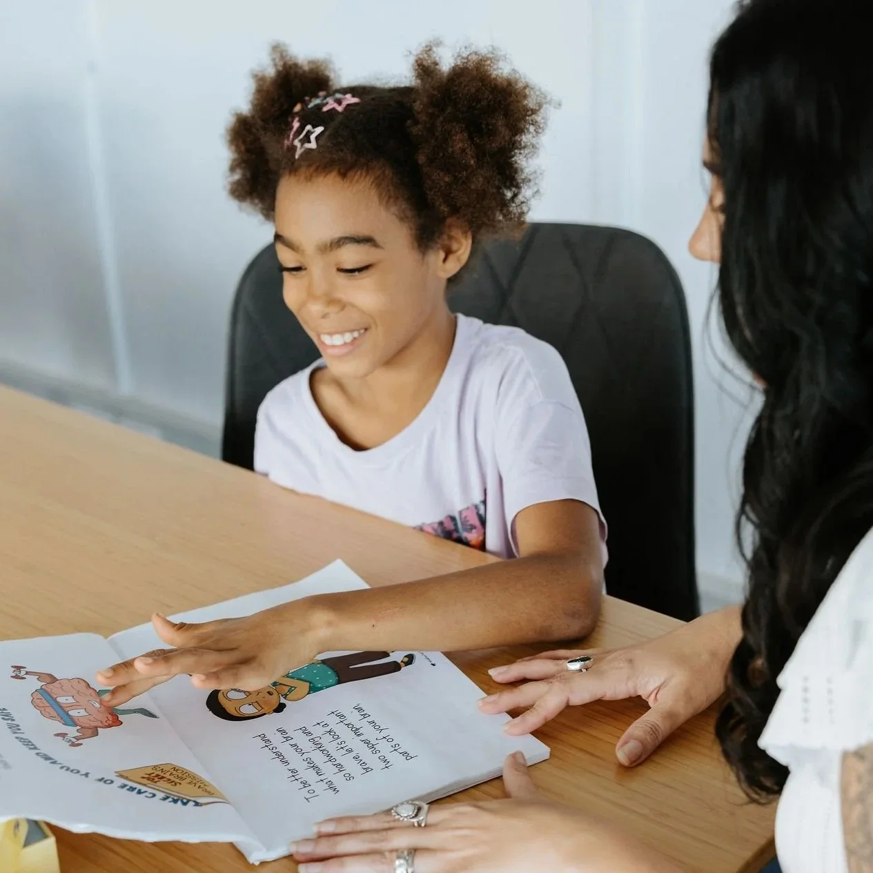 A PBS client is smiling and touching a colorful illustrated book on a wooden table. Candice Heffernan is sitting beside her, helping her with the book.
