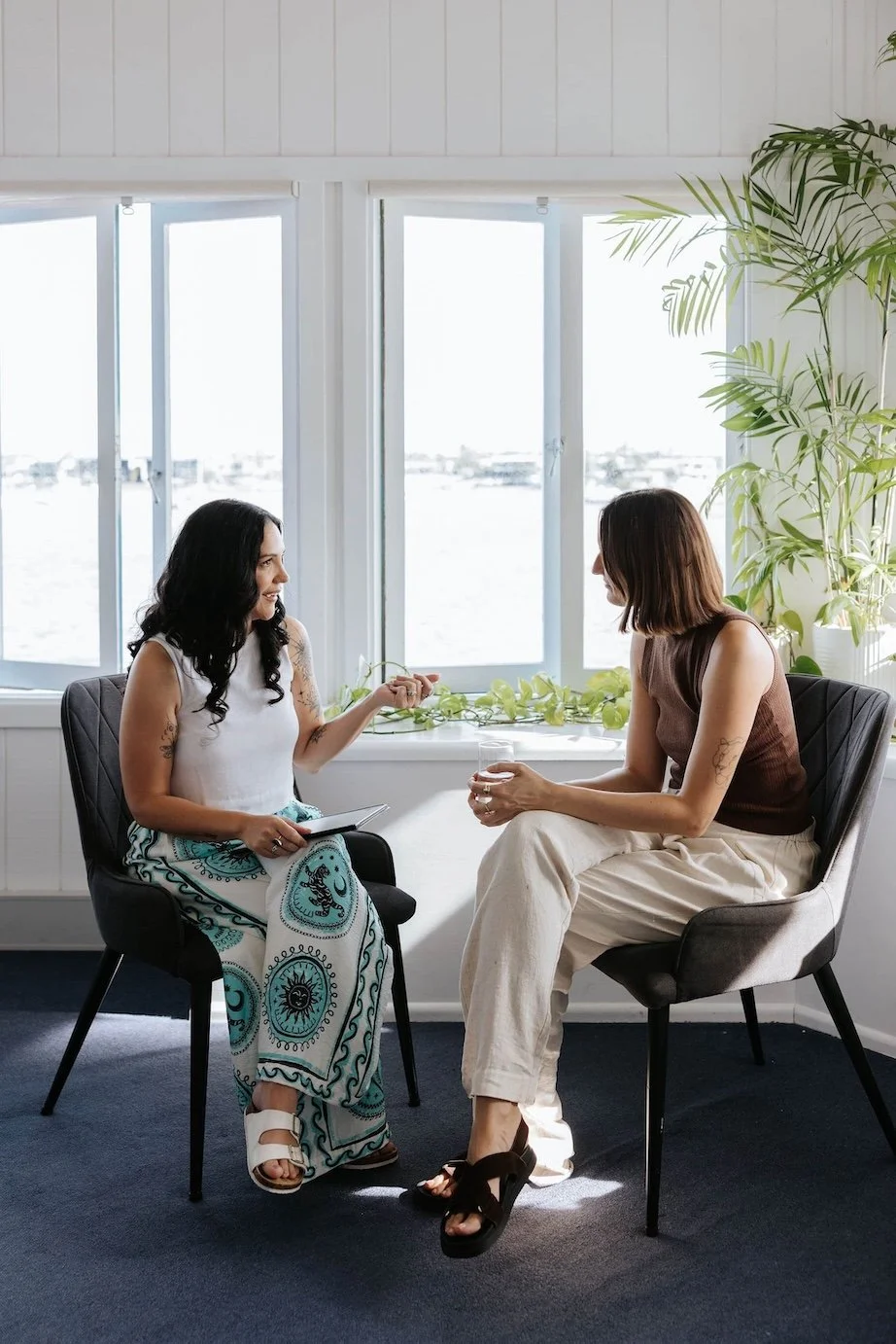 Candice Heffernan and a client sitting in chairs facing each other, engaged in conversation, with a window overlooking water in the background and a large green plant beside them.