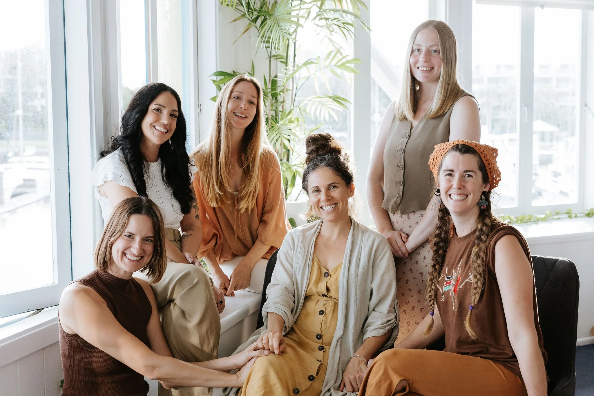 Group of smiling women gathered in a bright, indoor space with large windows and a potted plant in the background.
