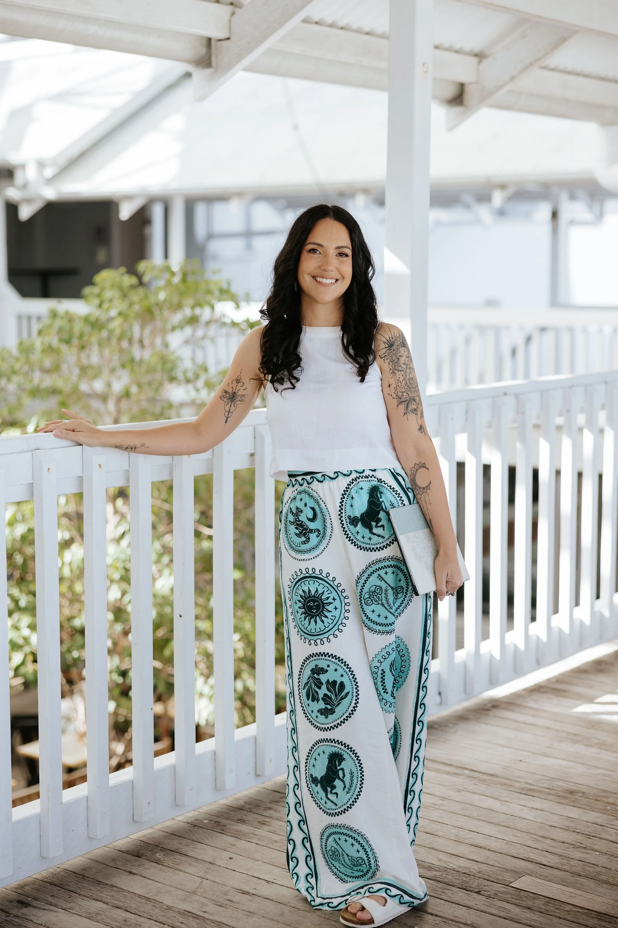 Candice Heffernan wearing a white sleeveless top and wide-leg pants with celestial designs, standing on a wooden porch with a white railing under a white roof. She holds a clipboard and stands near plants and a railing.
