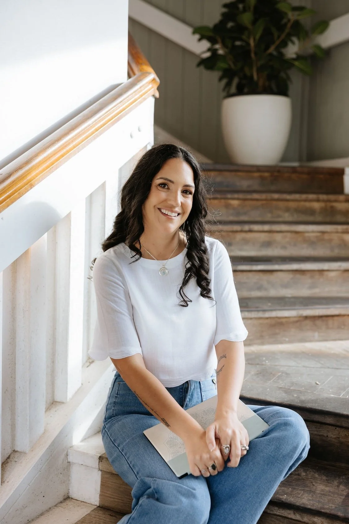 Candice Heffernan smiling, sitting on wooden stairs indoors, wearing a white top and blue jeans, holding a notebook.