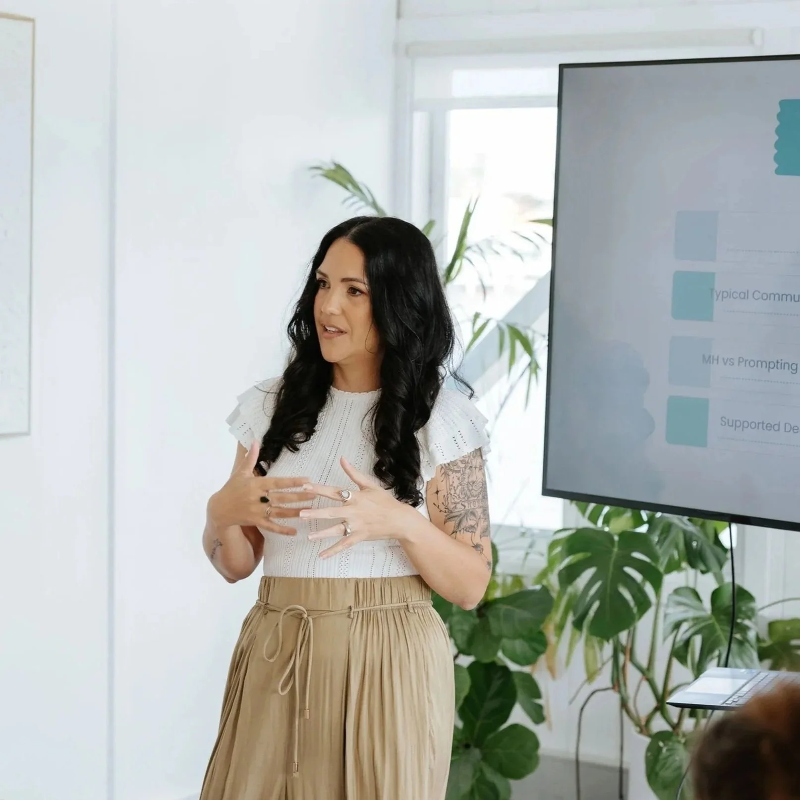 Candice Heffernan dressed in a white top and beige skirt, is giving a presentation for a client of Amity Behaviour Consultung, in a bright room with large windows and green plants, in front of a screen displaying a presentation slide.