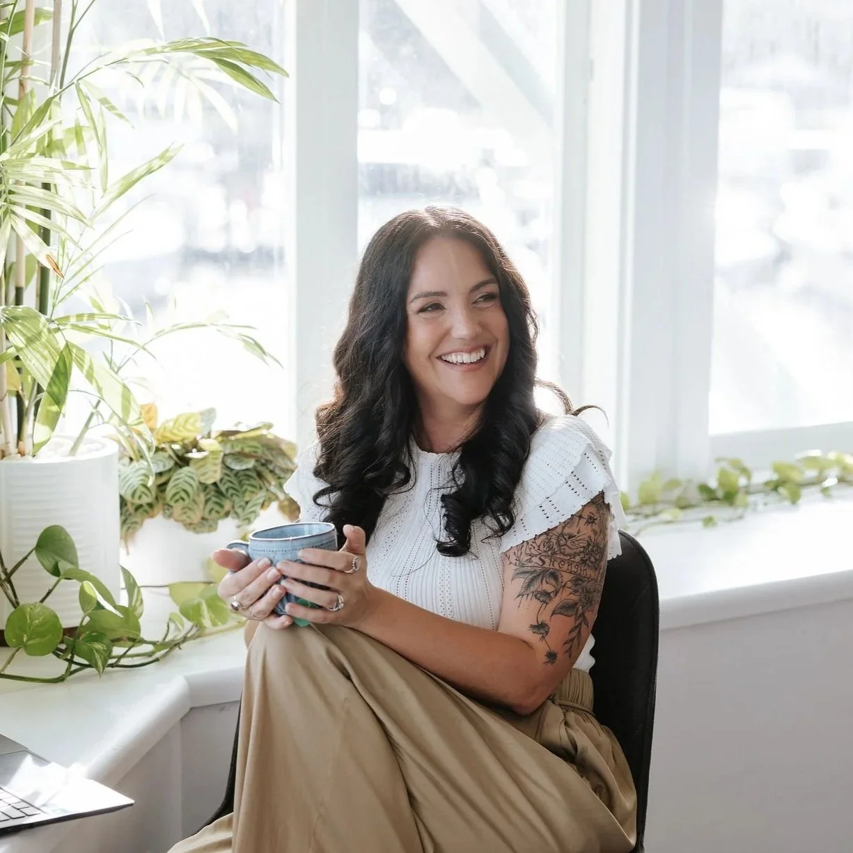 Candice Heffernan smiling and holding a blue mug, sitting in a bright room with large windows and potted plants.
