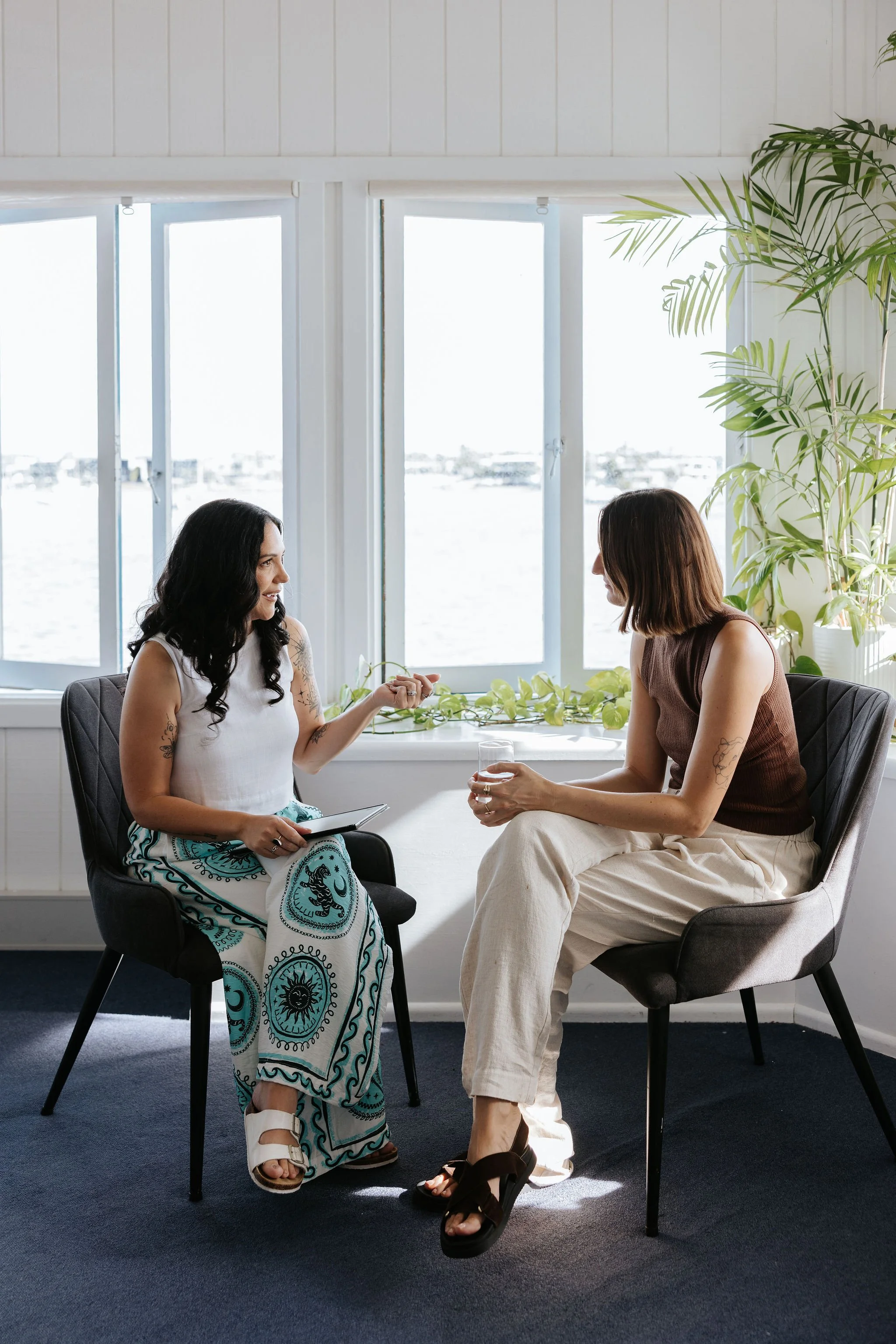 Candice Heffernan and a client sitting in chairs facing each other, engaged in conversation, with a window overlooking water in the background and a large green plant beside them.