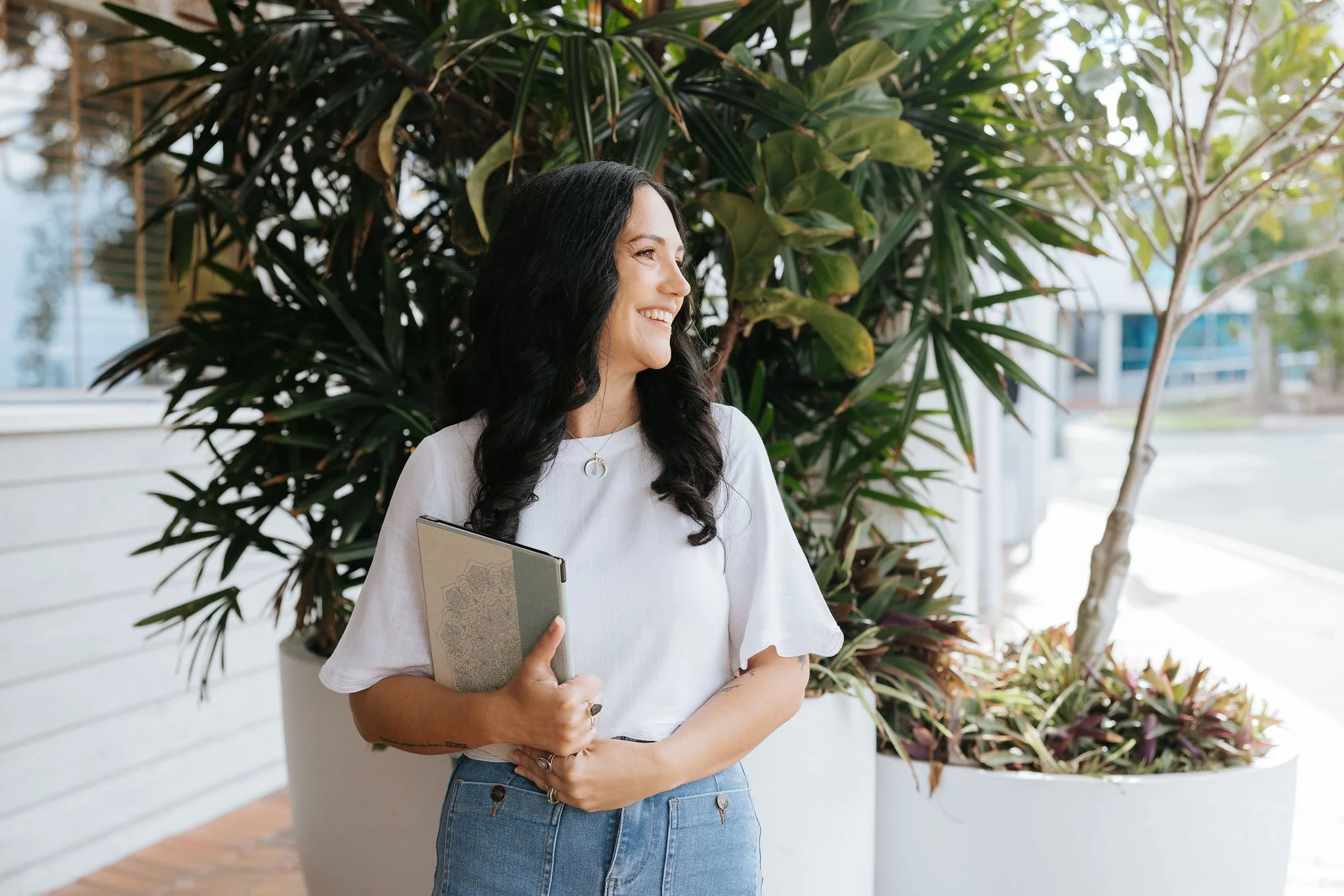 Candice Heffernan smiling, holding a notebook, standing outdoors next to large green potted plants.