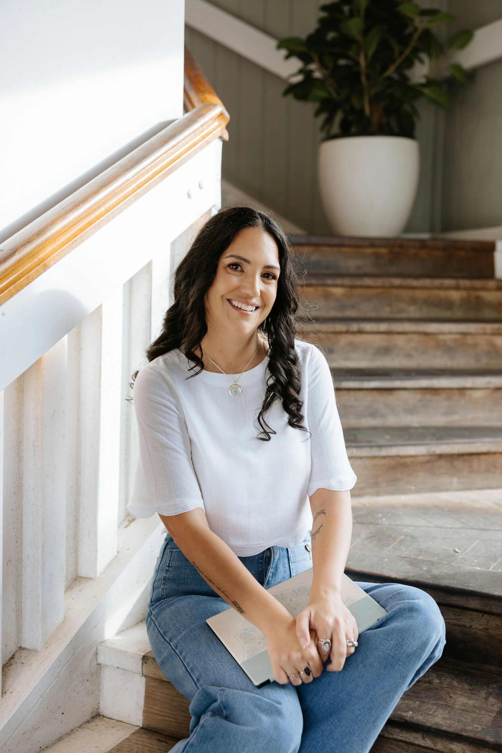 Candice Heffernan wearing a white top and blue jeans, smiles while sitting on wooden stairs indoors, holding a tablet in her lap. There is a large potted plant in the background.