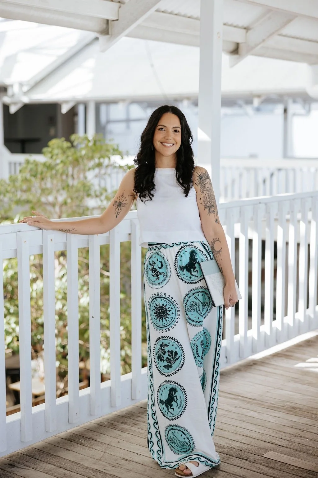 Candice Heffernan wearing a white sleeveless top and wide-leg pants with celestial designs, standing on a wooden porch with a white railing under a white roof. She holds a clipboard and stands near plants and a railing.