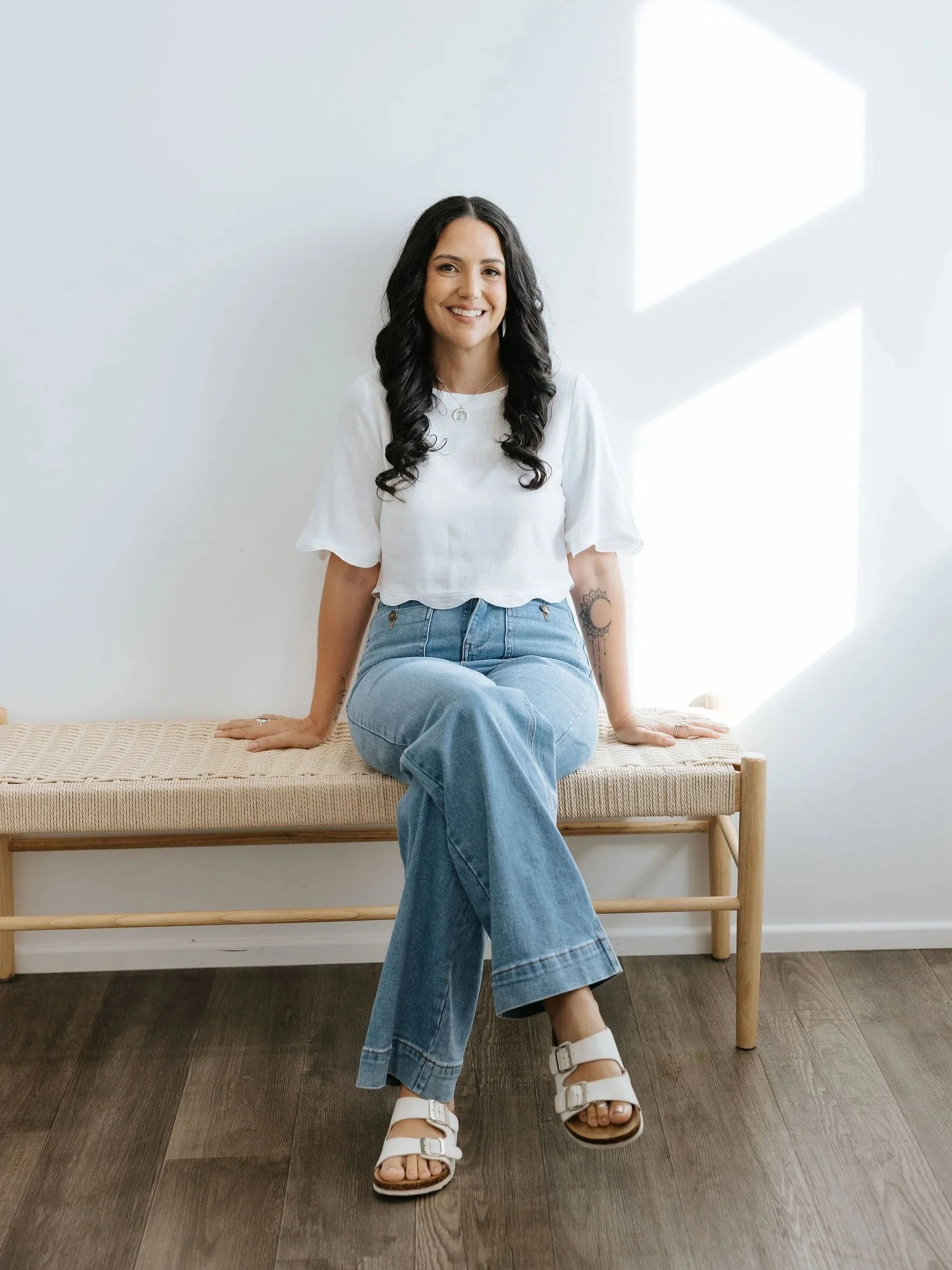 Candice Heffernan is sitting on a wooden bench in a bright room with white walls and wooden floor. She is wearing a white short-sleeved top, blue jeans, and white sandals. She is smiling and looking at the camera