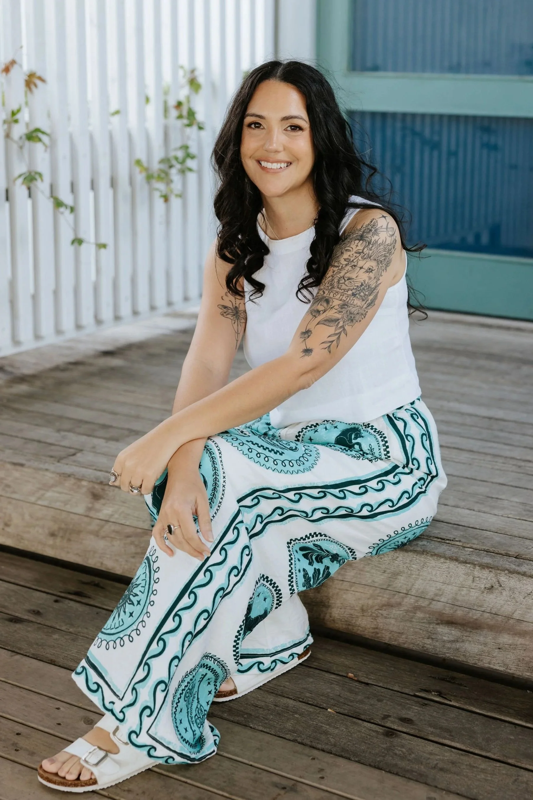 Candice Heffernan sitting on wooden decking, wearing a white sleeveless top and a patterned blue and white pants, with white sandals.