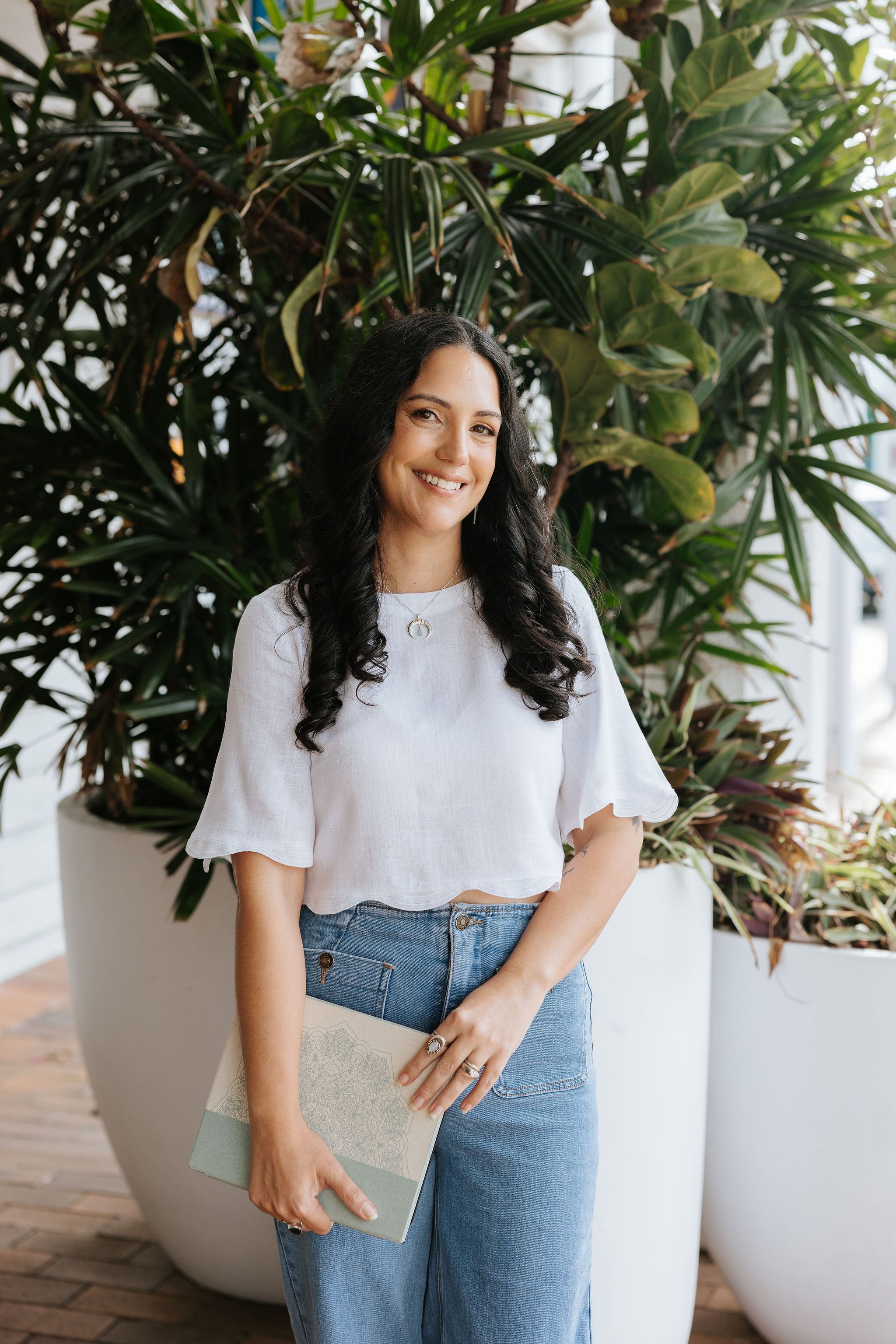 Candice Heffernan stands in front of large green plants in a well-lit indoor setting. She is wearing a white ruffled blouse and blue jeans, holding a notebook with a detailed cover.