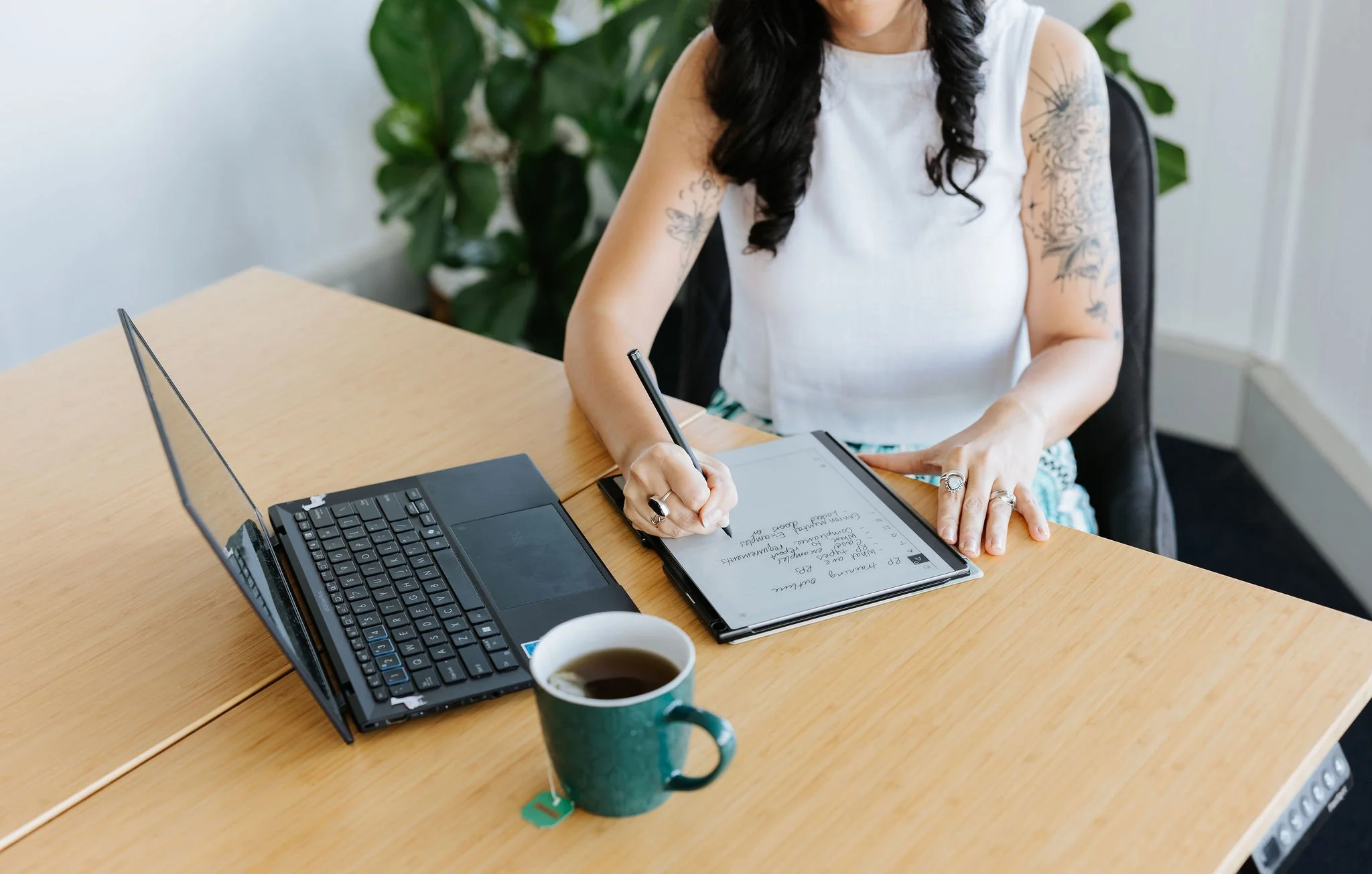 Candice Heffernan sitting at a wooden desk. She is writing on a tablet with a stylus. There is an open laptop and a cup of coffee on the desk, and green plants are in the background.