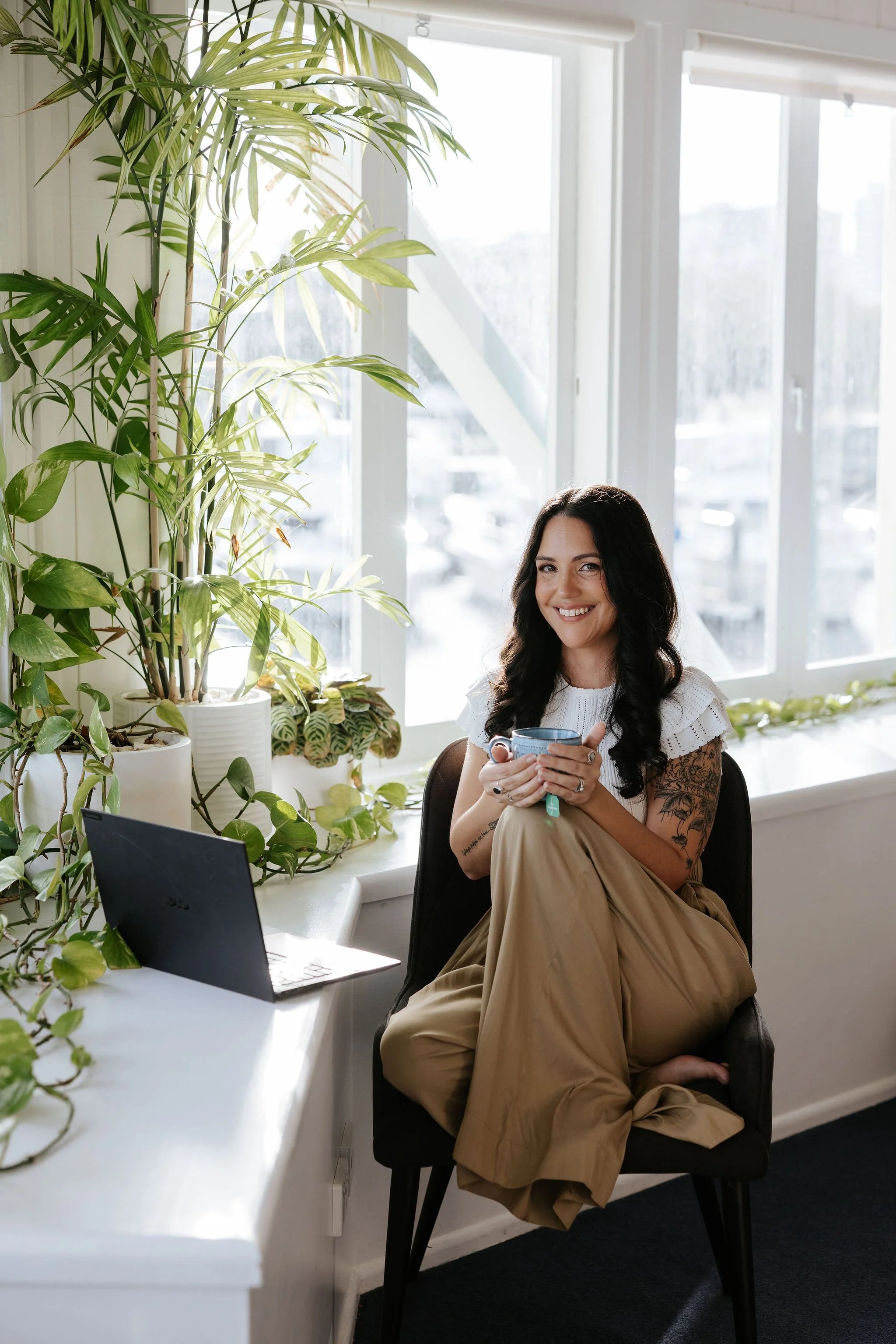 Candice Heffernan smiling and sitting cross-legged on a black chair in an office. The office has large windows with blinds and is decorated with indoor plants on a white desk and windowsill.