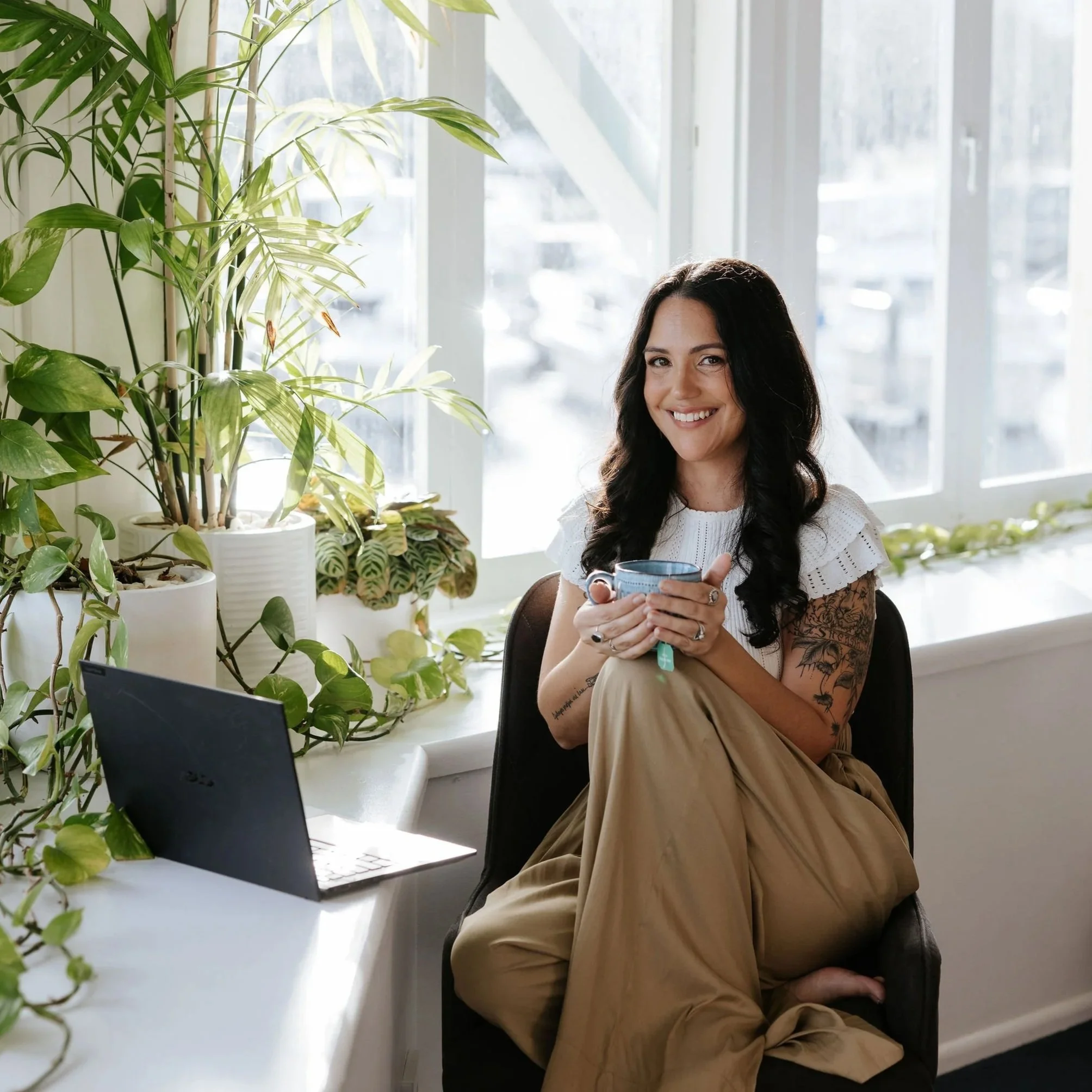 Candice Heffernan sitting in front of her computer in a sunlit room filled with green potted plants, holding a mug.