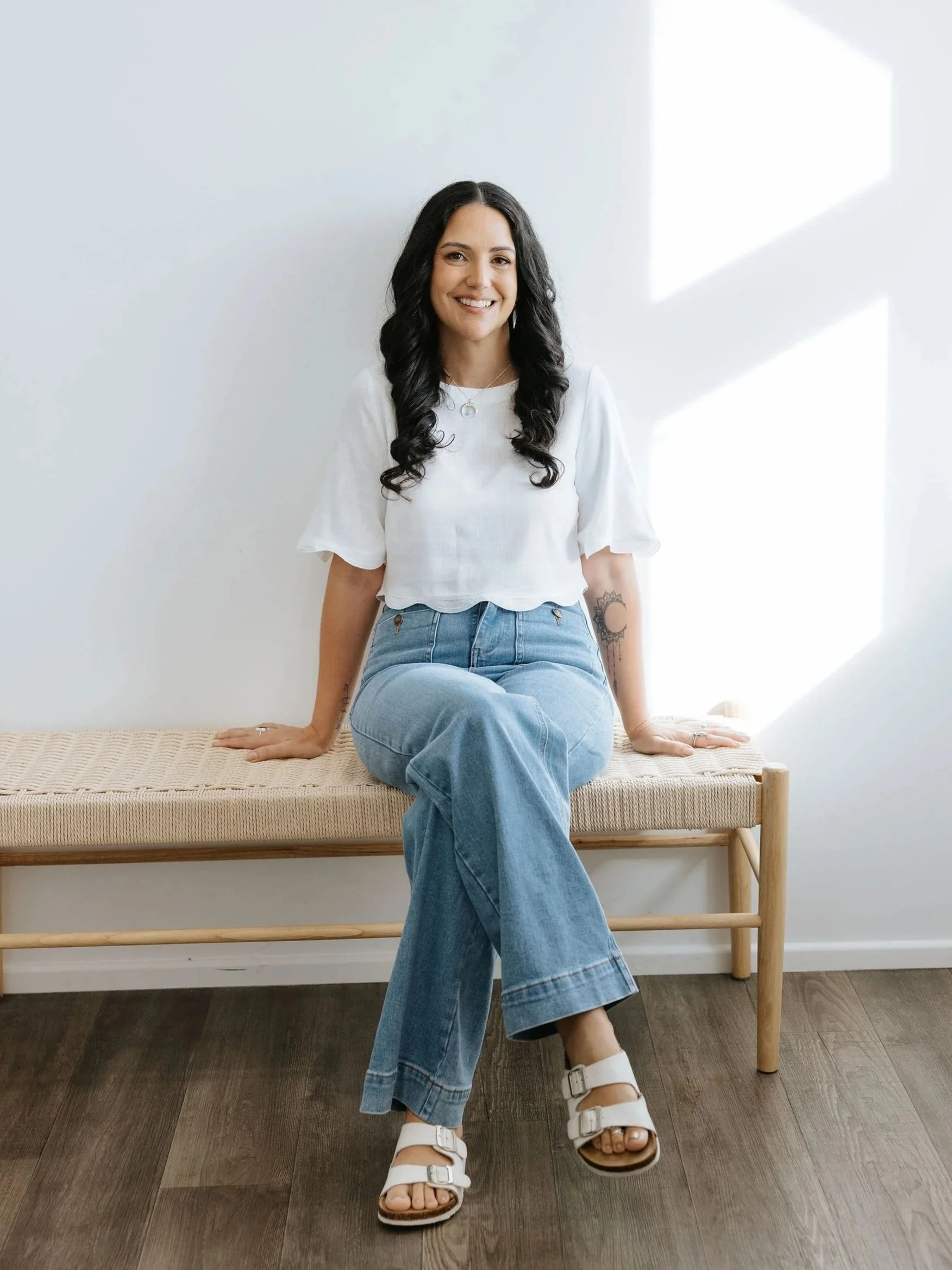 Candice Heffernan is sitting on a wooden bench in a bright room with white walls and wooden floor. She is wearing a white short-sleeved top, blue jeans, and white sandals. She is smiling and looking at the camera