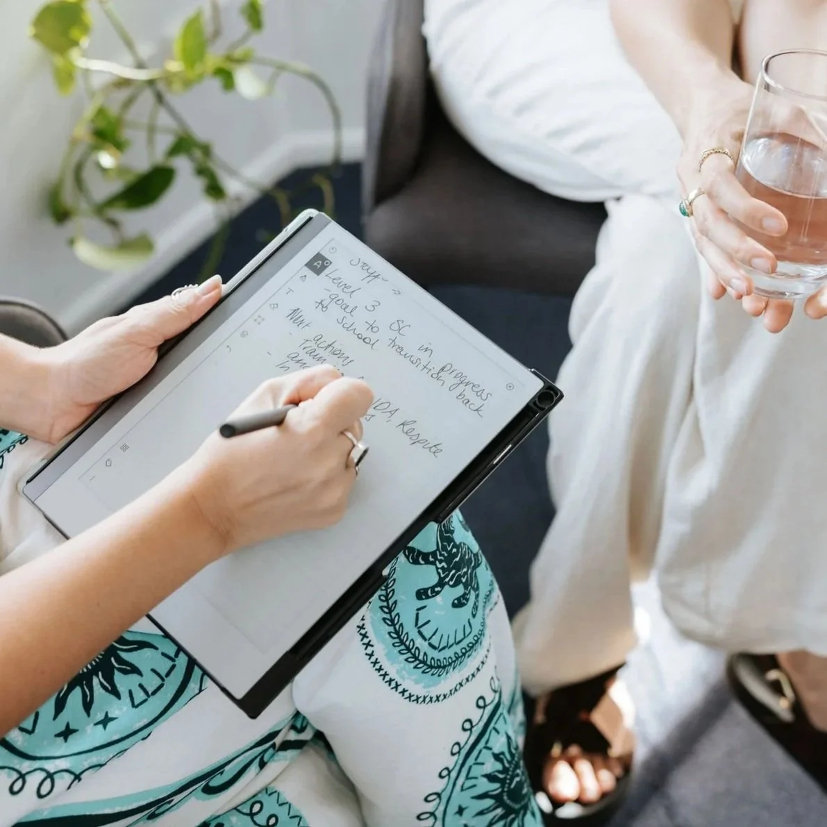 Candice Heffernan holding a stylus and writing notes on a tablet, with another person standing nearby holding a glass of water. The notes on the tablet include references to school and activities.