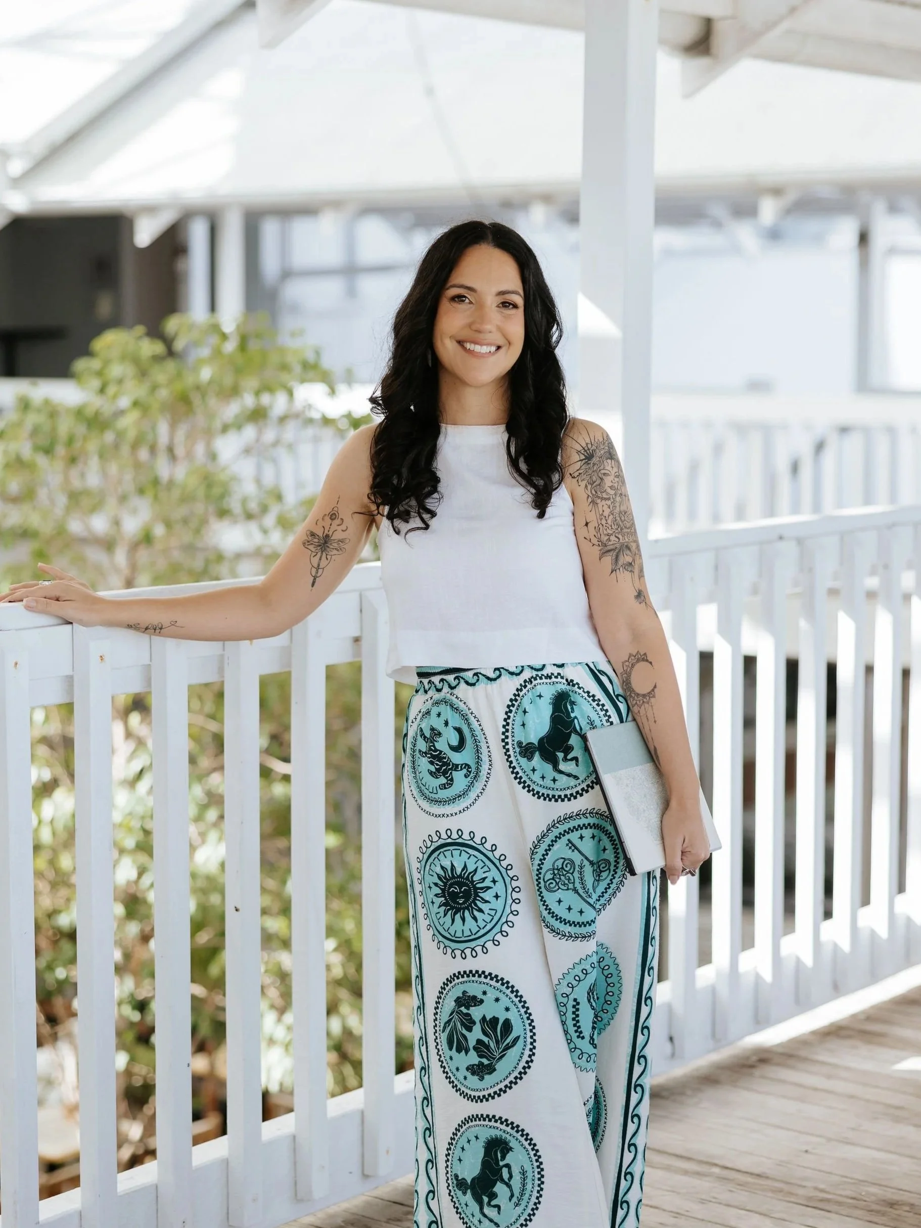 Candice Heffernan, smiling and standing on a wooden porch with a white railing, holding a tablet in her left hand, wearing a white sleeveless top and wide-leg pants with a blue and black zodiac pattern.