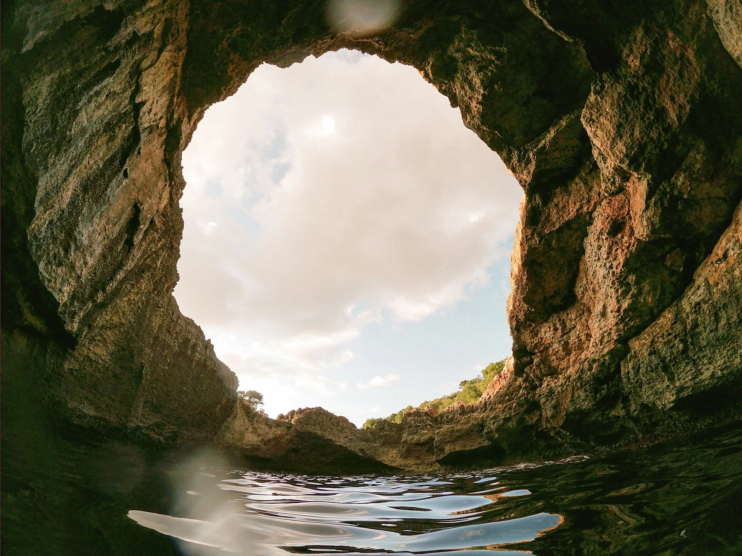 View from inside a cave looking out at the sky with clouds and sunlight, surrounded by rocky walls.