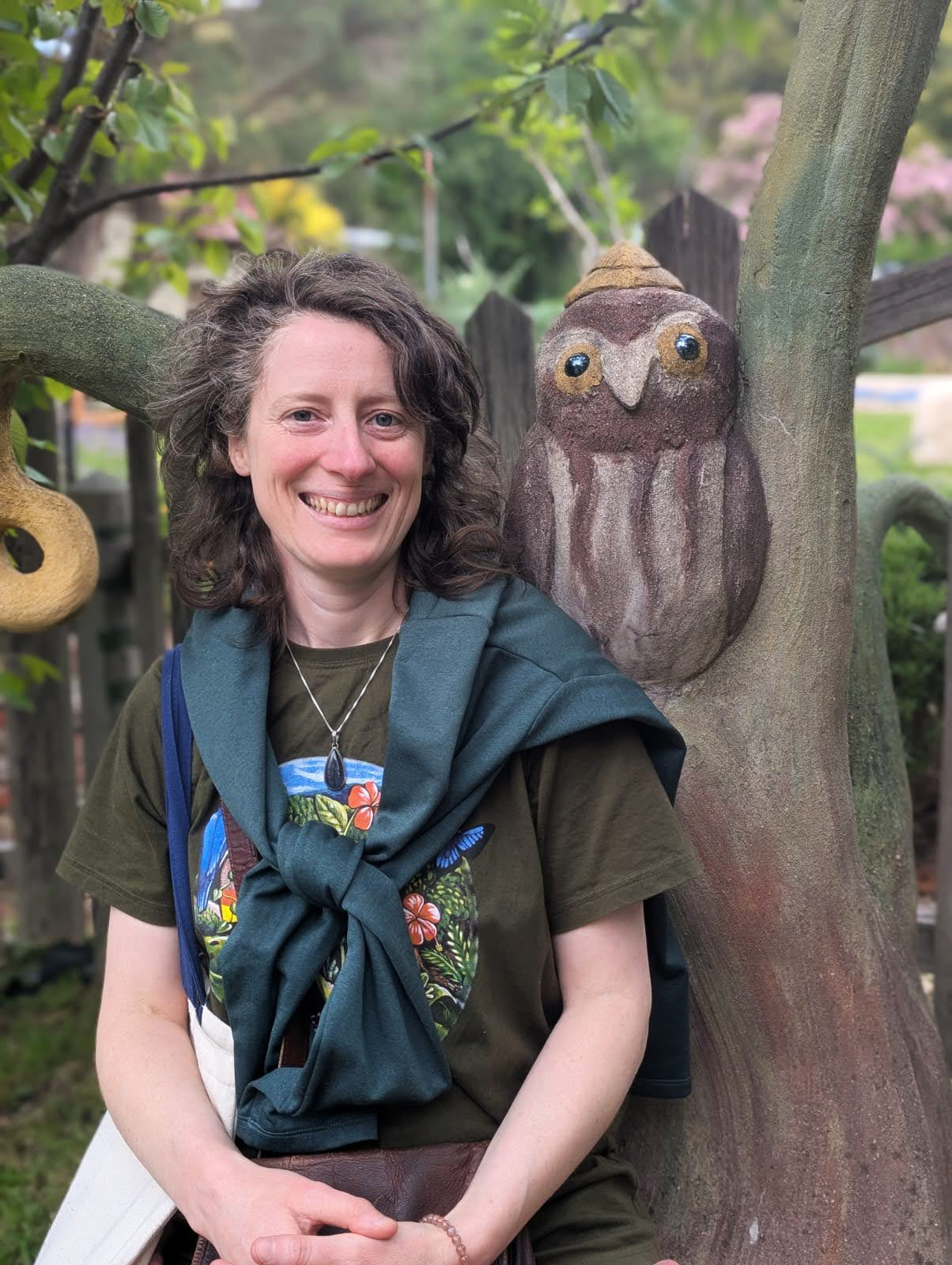 A woman with curly brown hair smiling, wearing a green shirt with a colorful design, a necklace, and a blue jacket tied around her shoulders, sitting outdoors near a tree with a painted owl sculpture on it.