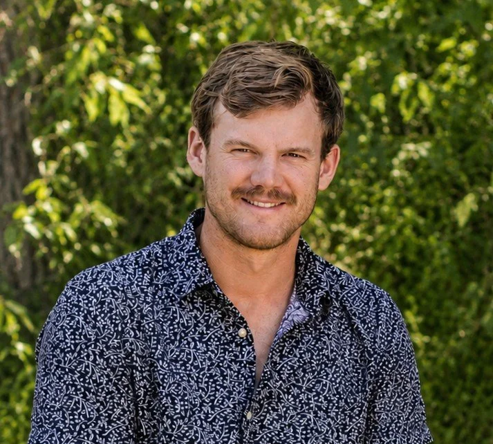 A smiling man with brown hair and a beard, wearing a patterned button-up shirt, standing outdoors with green foliage in the background. Brayden Fair