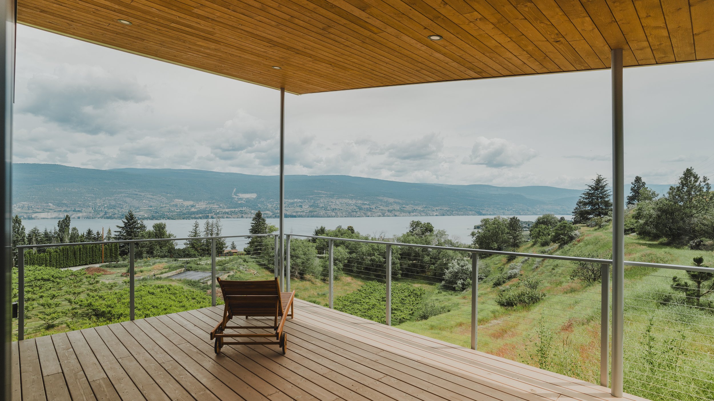 A spacious balcony with wooden flooring and a wooden ceiling, overlooking a landscape with trees, a body of water, and distant mountains, with a wooden lounge chair on the balcony.