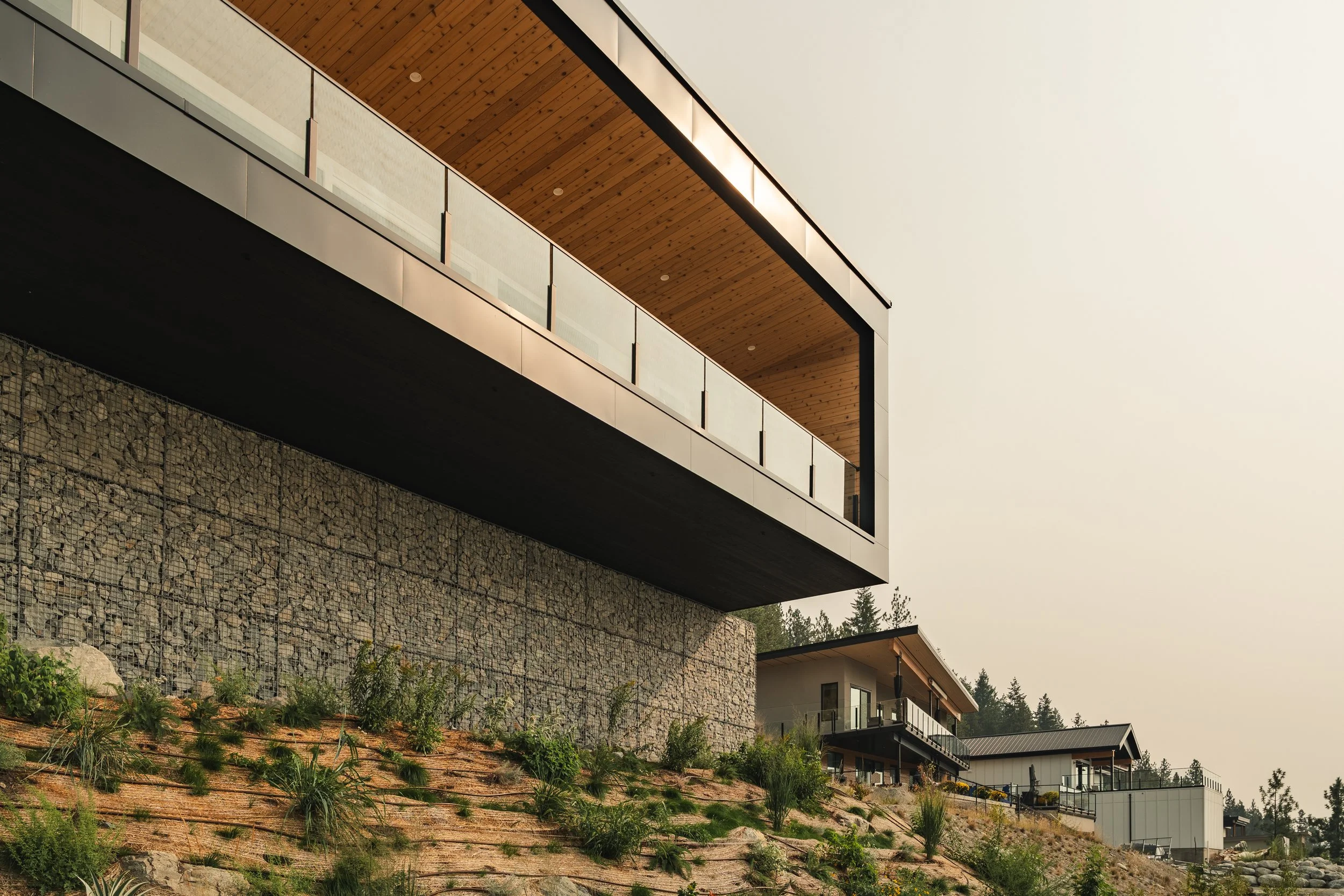Modern house with wood, glass, and metal materials on a hillside with landscaped garden and neighboring homes, under an overcast sky. Naramata BC