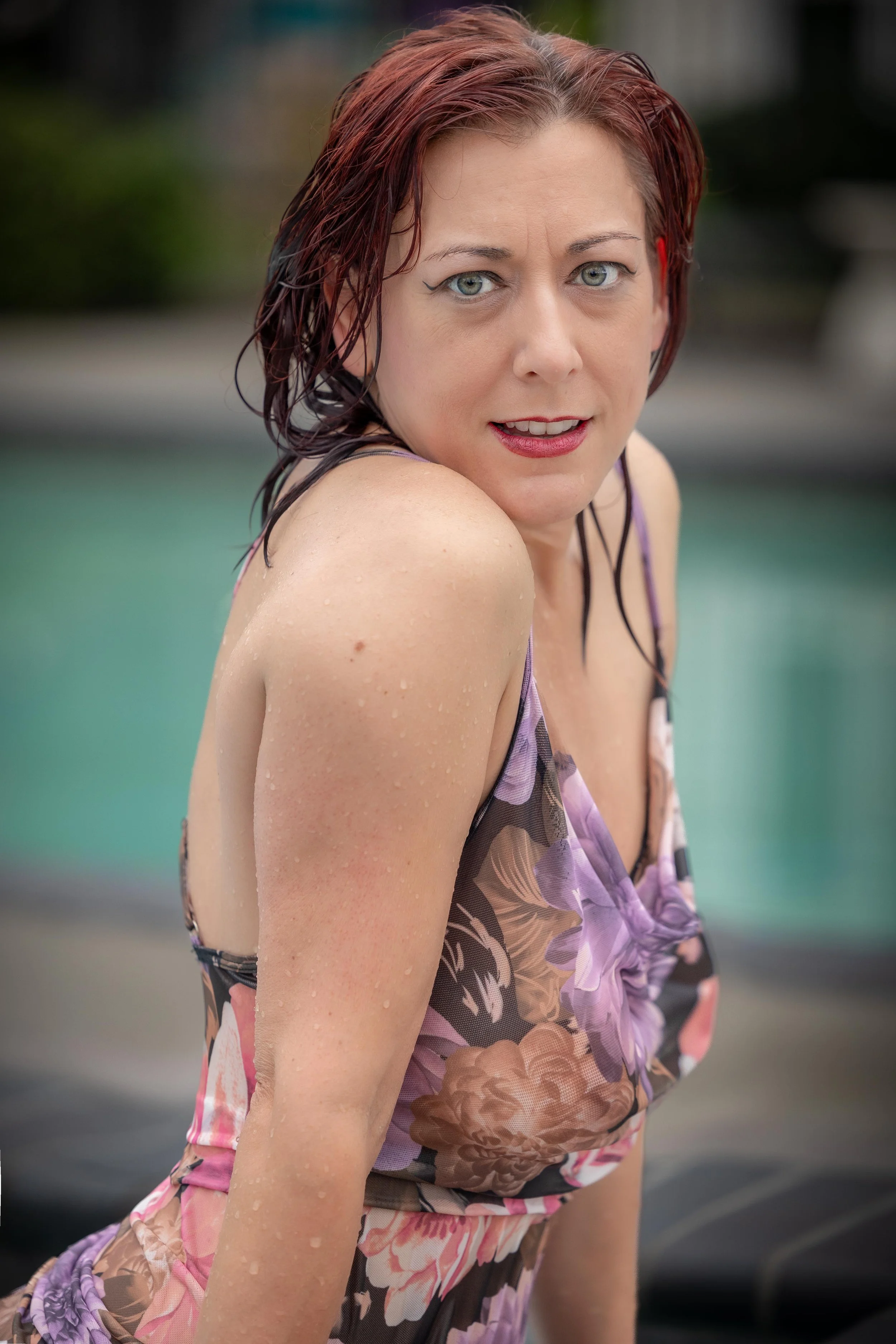 Close-up of a woman with wet, reddish-brown hair and light eyes, wearing a floral dress, outdoors near a swimming pool.