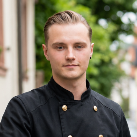 Ein junger Mann trägt eine schwarze Schornsteinfegerjacke mit goldenen Knöpfen, steht vor einem grünen, bewaldeten Hintergrund.