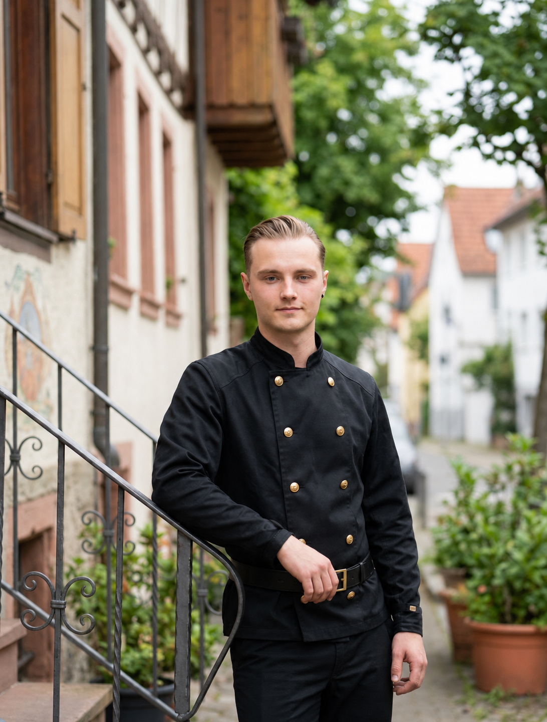 Ein junger Mann in einer schwarzen Schornsteinfegerjacke mit goldenen Knöpfen steht vor einer alten Häuserwand im Freien.