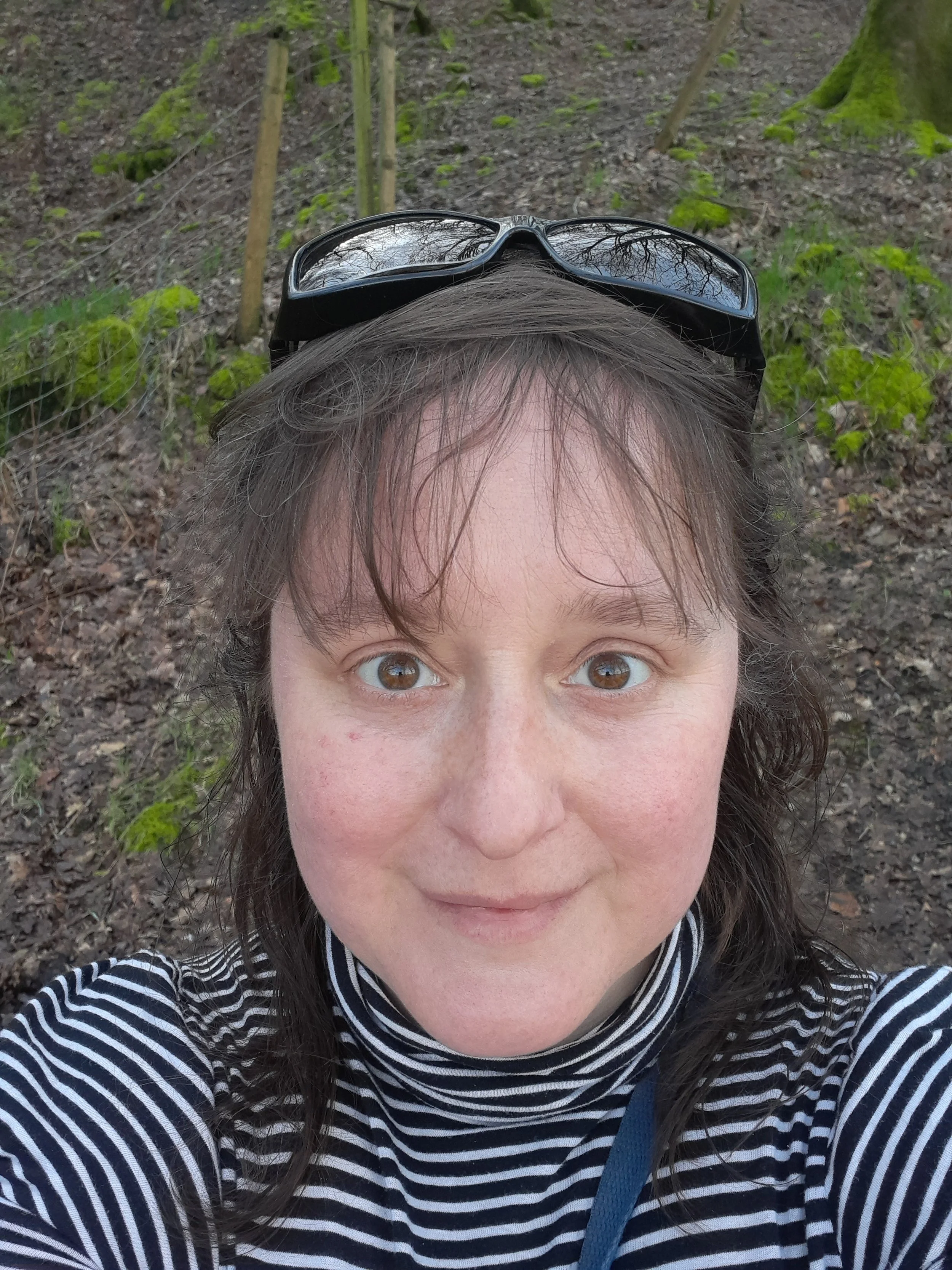 Close-up of a smiling woman with brown eyes and brown hair taking a selfie outdoors. She is wearing sunglasses on her head and a black and white striped shirt. The background shows a wooded area with trees and green plants.