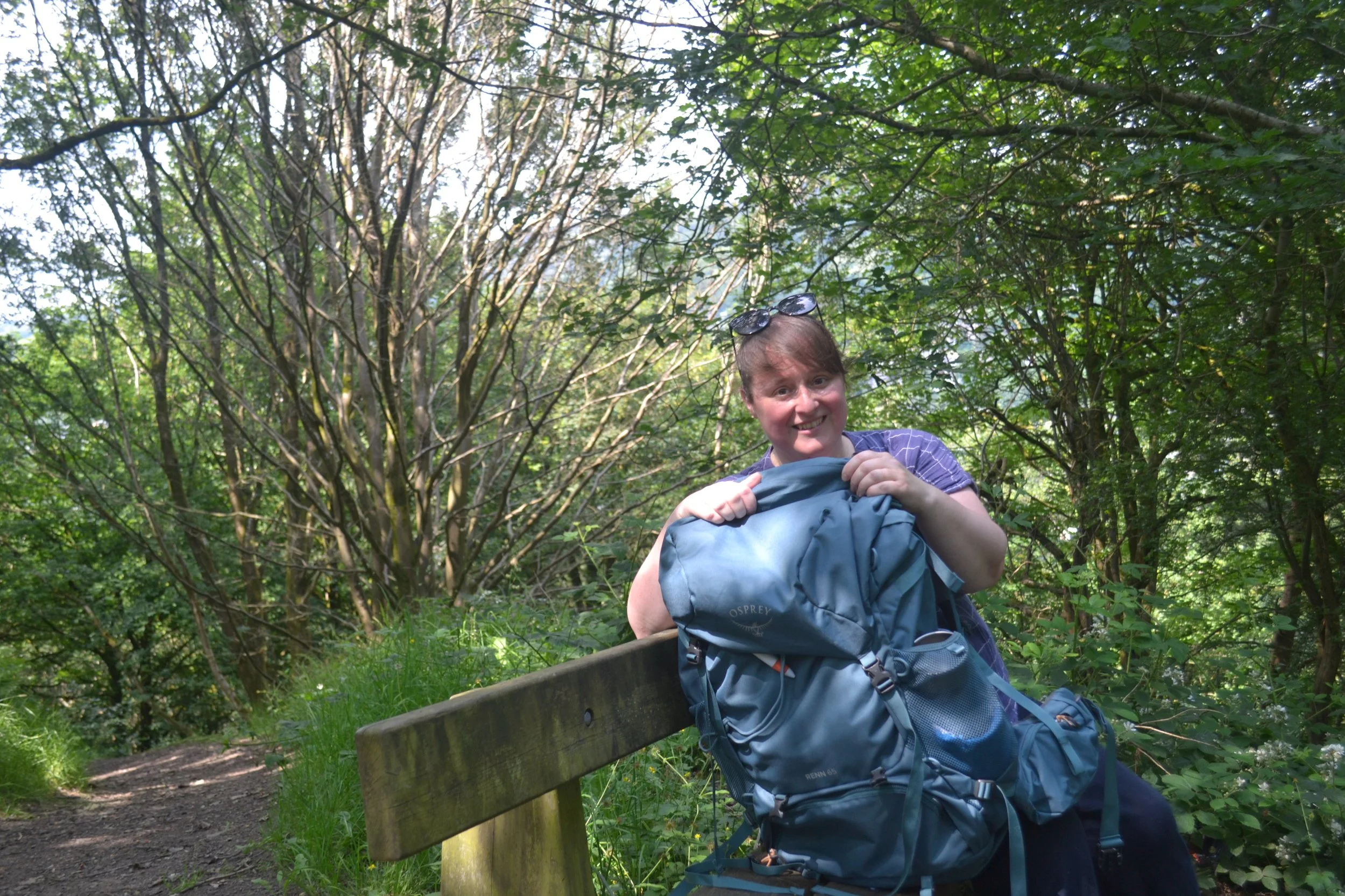 A smiling woman with a large rucksack sat on a bench in woodland