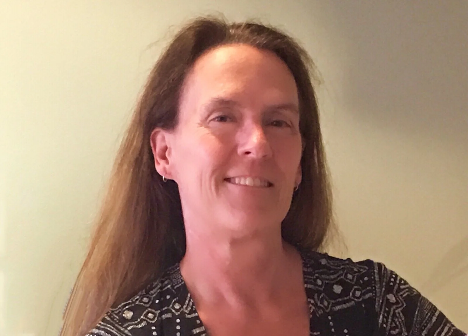 A woman with long brown hair, smiling, wearing a patterned black and white top, standing against a plain light yellow wall.