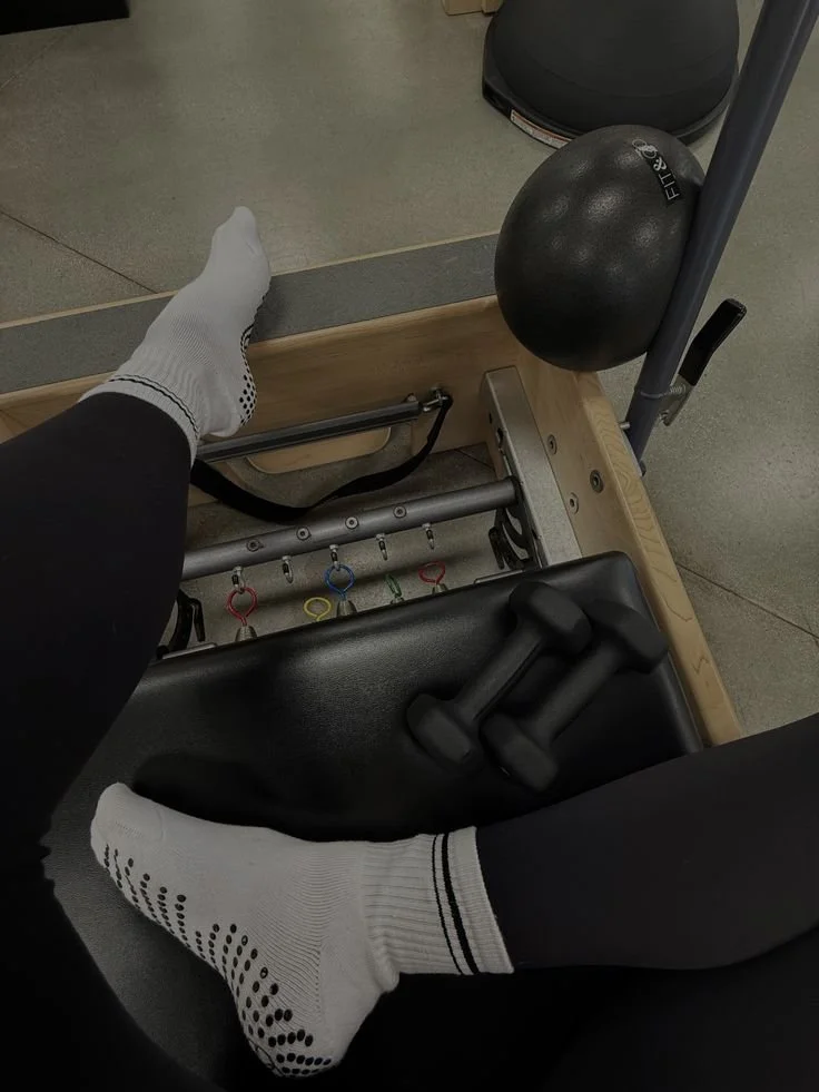 View from above of a person with white socks and black leggings sitting at a physical therapy or rehabilitation gym machine, with a black rubber ball, ankle weights, and resistance bands visible.